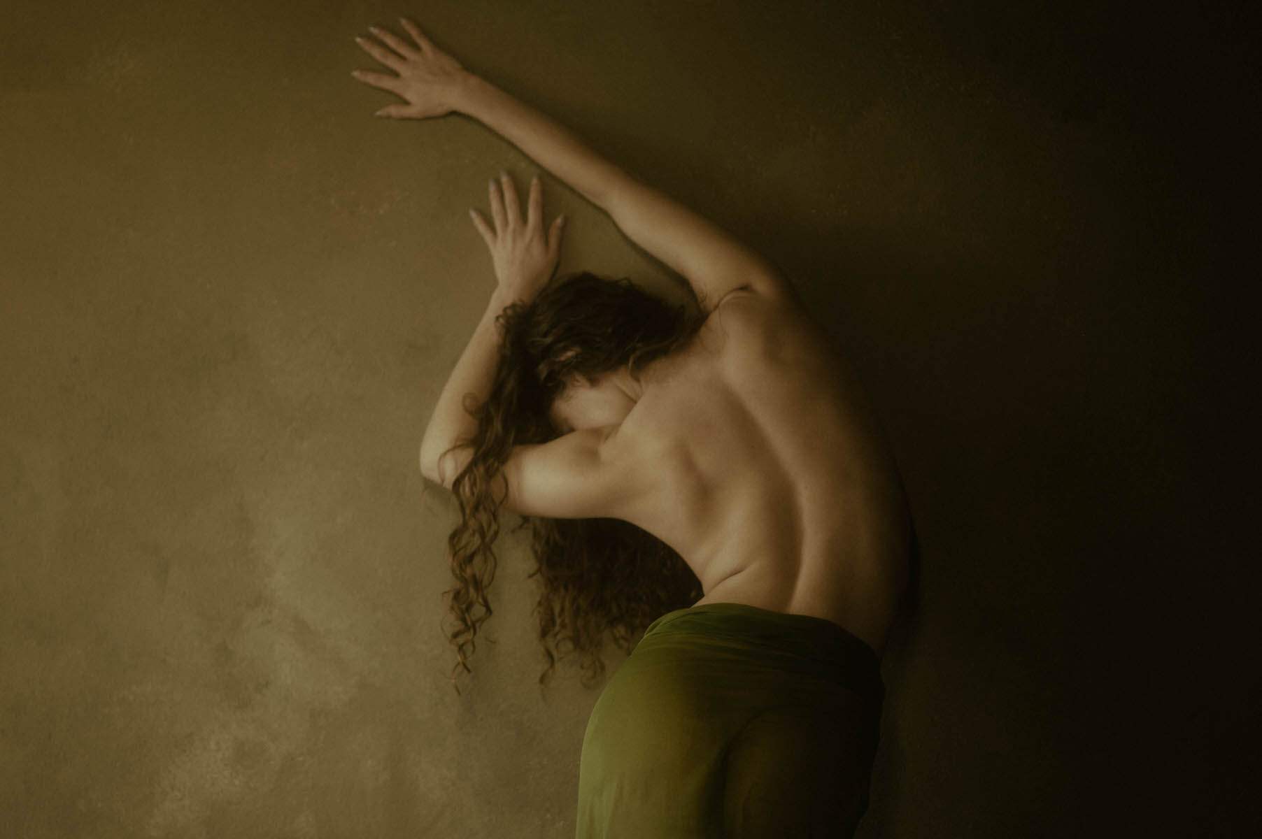 A woman with long hair, draped in a green cloth, leans forward with their back facing the camera and arms extended against a wall, capturing the essence of a fine art boudoir.