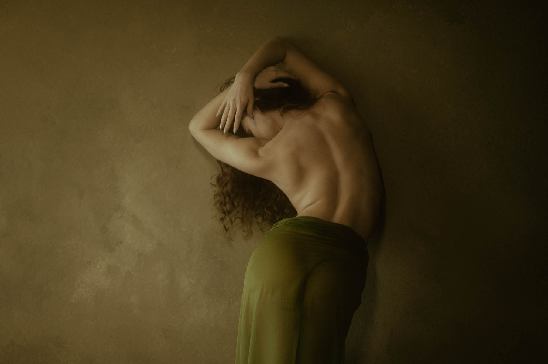 A topless woman with curly hair stands against a textured beige wall, their back to the camera, arms raised and head tilted. In this fine art boudoir setting, they are draped in a green cloth around the waist.