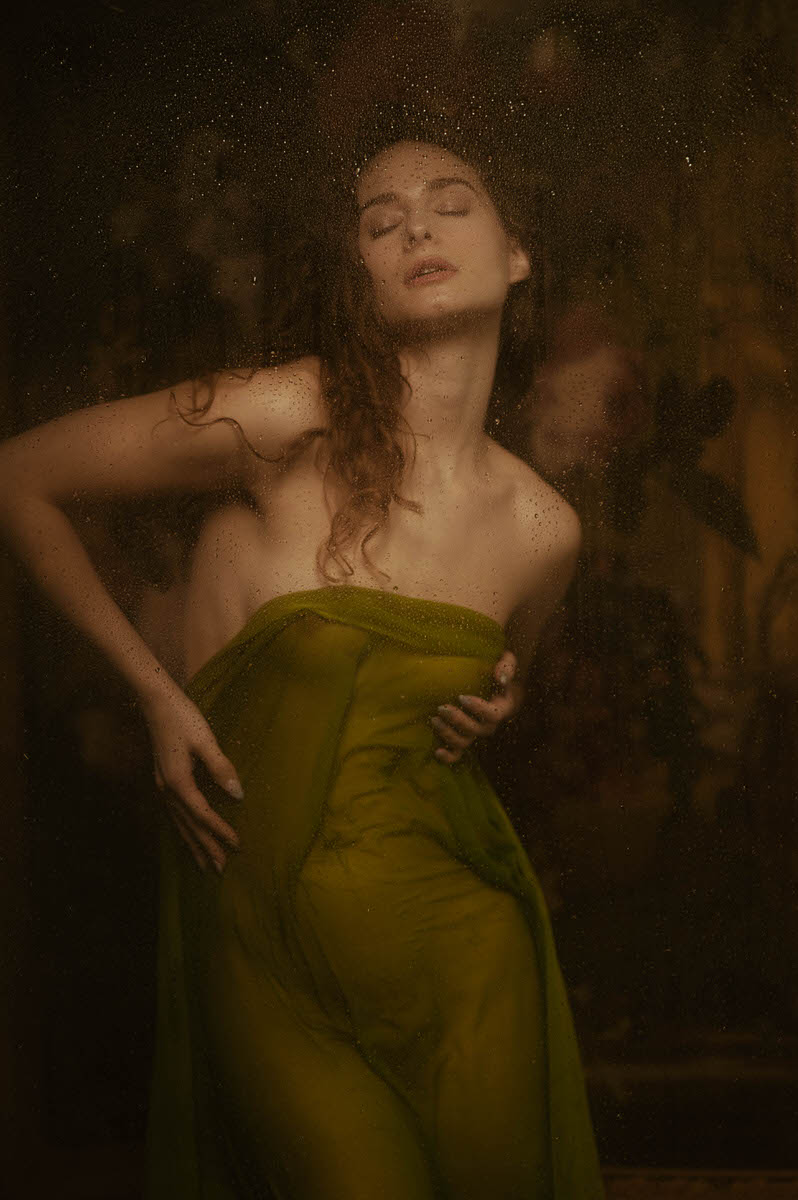 A woman with long curly hair poses behind a misty glass, draped in a green wet cloth, capturing a statuesque look for a fine art boudoir session in Dallas, TX.