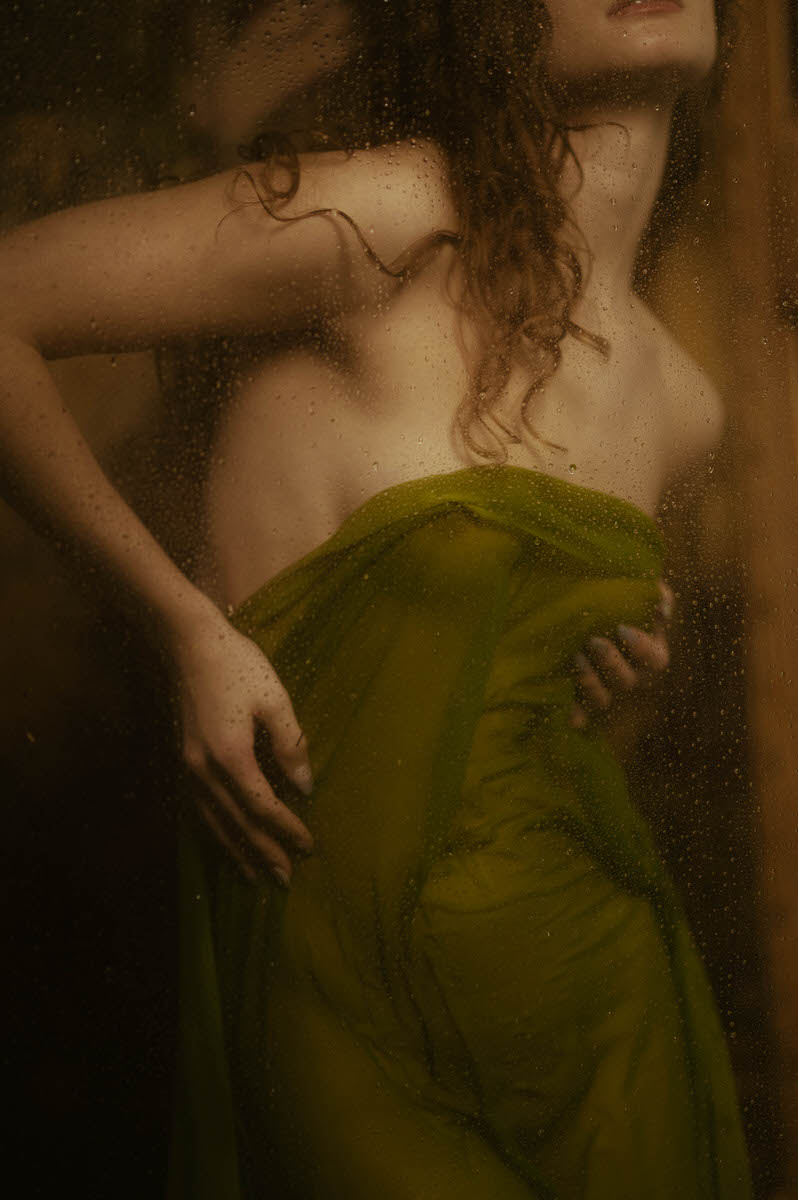 A person with wet hair is wrapped in wet green fabric behind a rain-splattered glass surface, evoking the mystique of fine art boudoir.