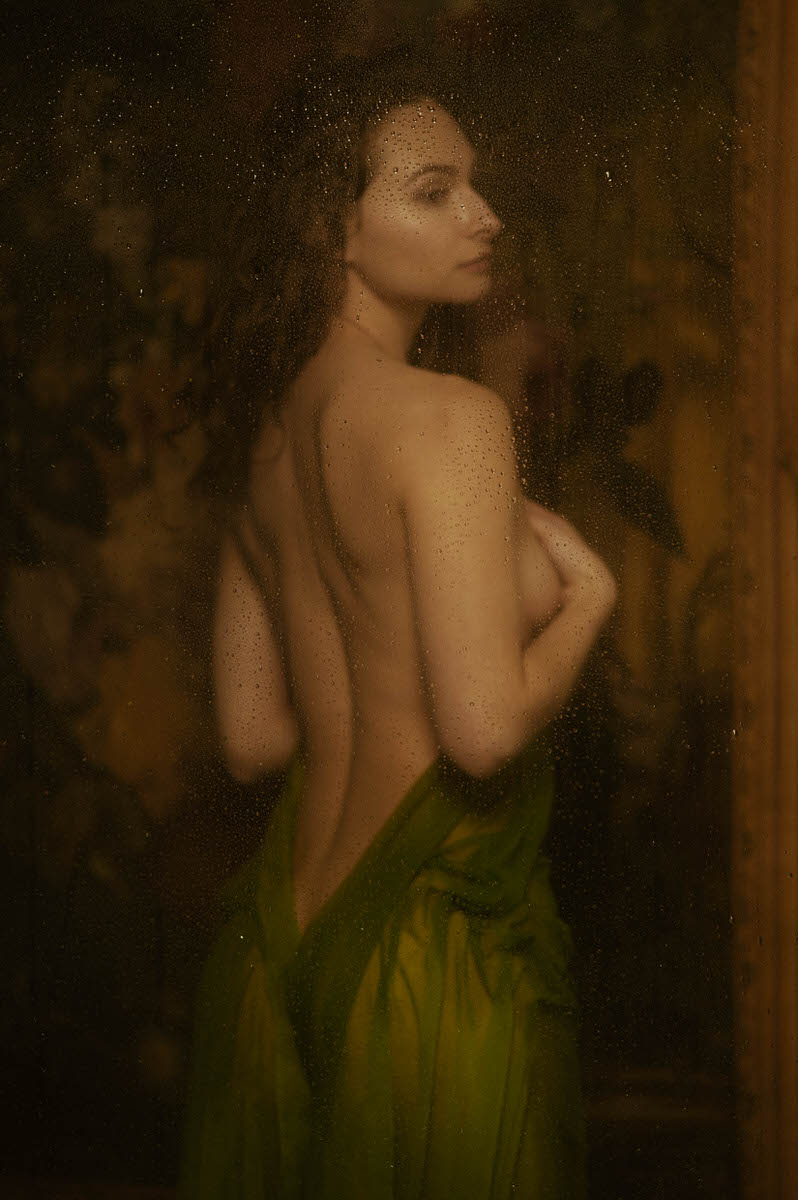 A woman with long dark hair, draped in wet green fabric, stands with their back partially turned, viewed through a misty, water-speckled surface, capturing the essence of a faux shower boudoir setup in Dallas, TX.