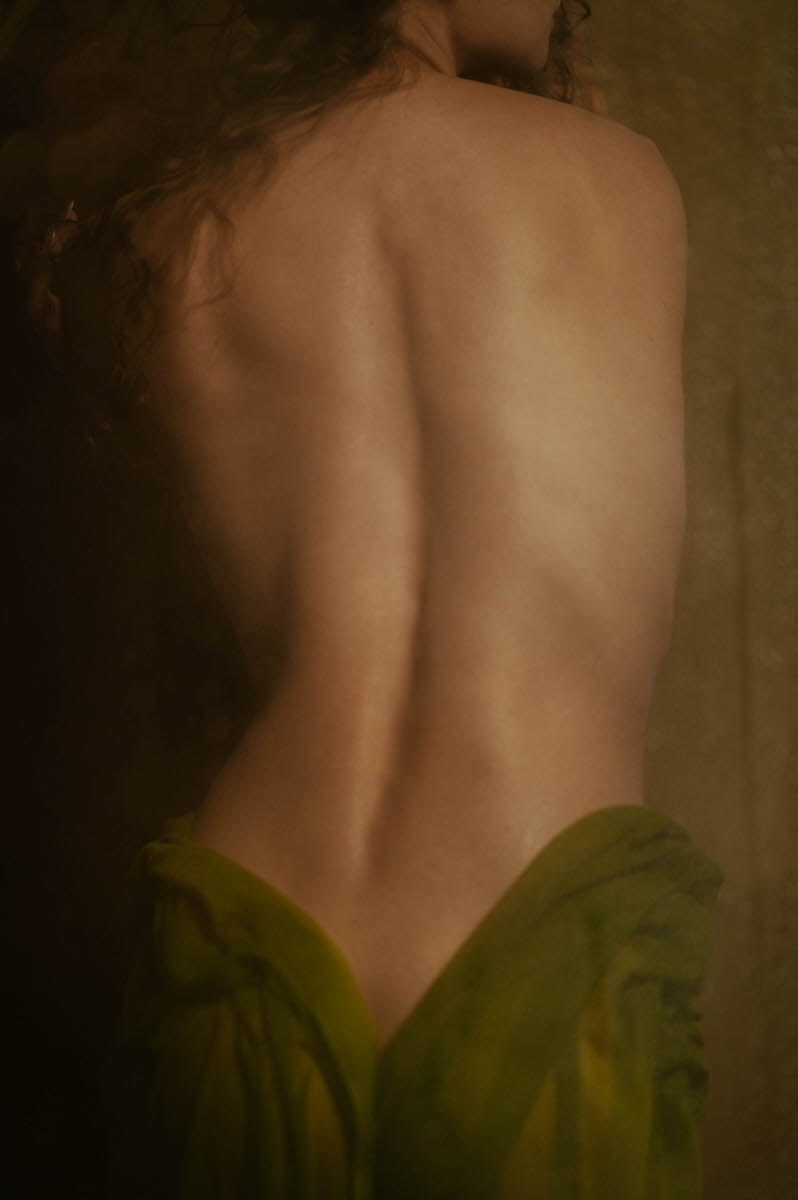 A woman with long curly hair stands with their back facing the camera, draped in a sheer wet green fabric around the waist, creating an ethereal fine art boudoir aesthetic in Dallas, TX.