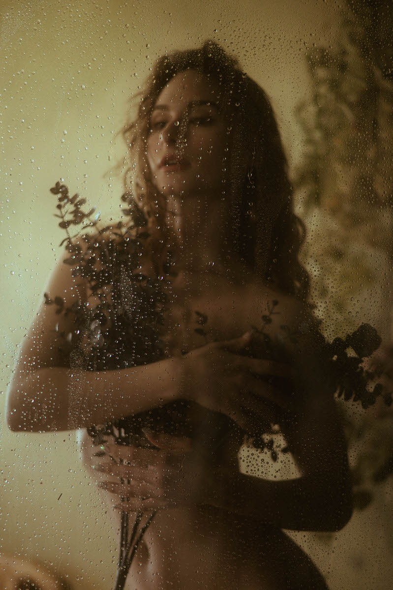 A person stands behind a rain-frosted glass, holding eucalyptus branches close to their chest, evoking the ethereal elegance of fine art boudoir photography with a blurred background.