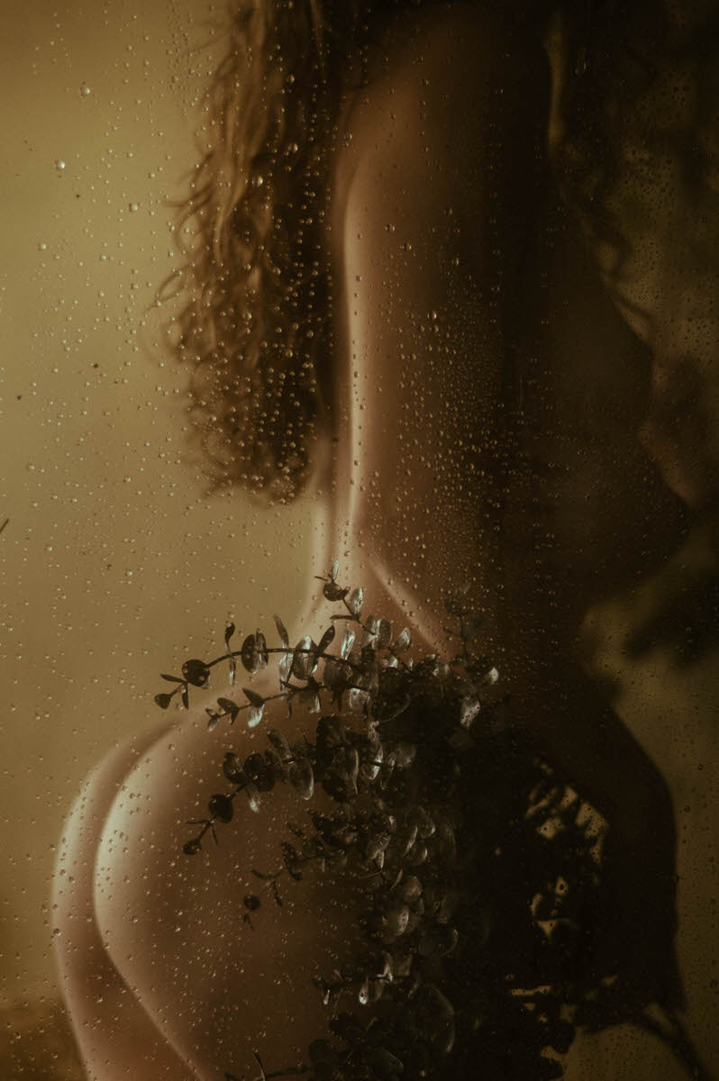 A silhouette of a nude woman with long curly hair holding a bouquet of dried flowers, seen through a wet glass surface, embodies the ethereal beauty of fine art nude photography.