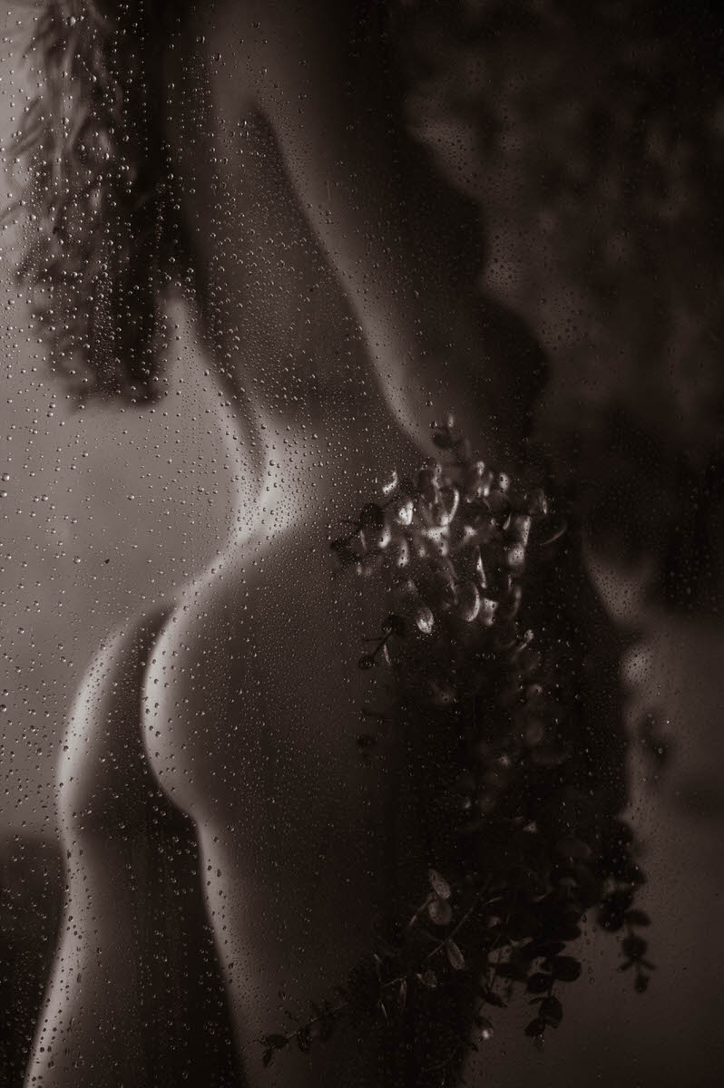 A fine art nude photograph featuring a silhouette of a person standing behind rain-covered glass, with water droplets accentuating the surface.