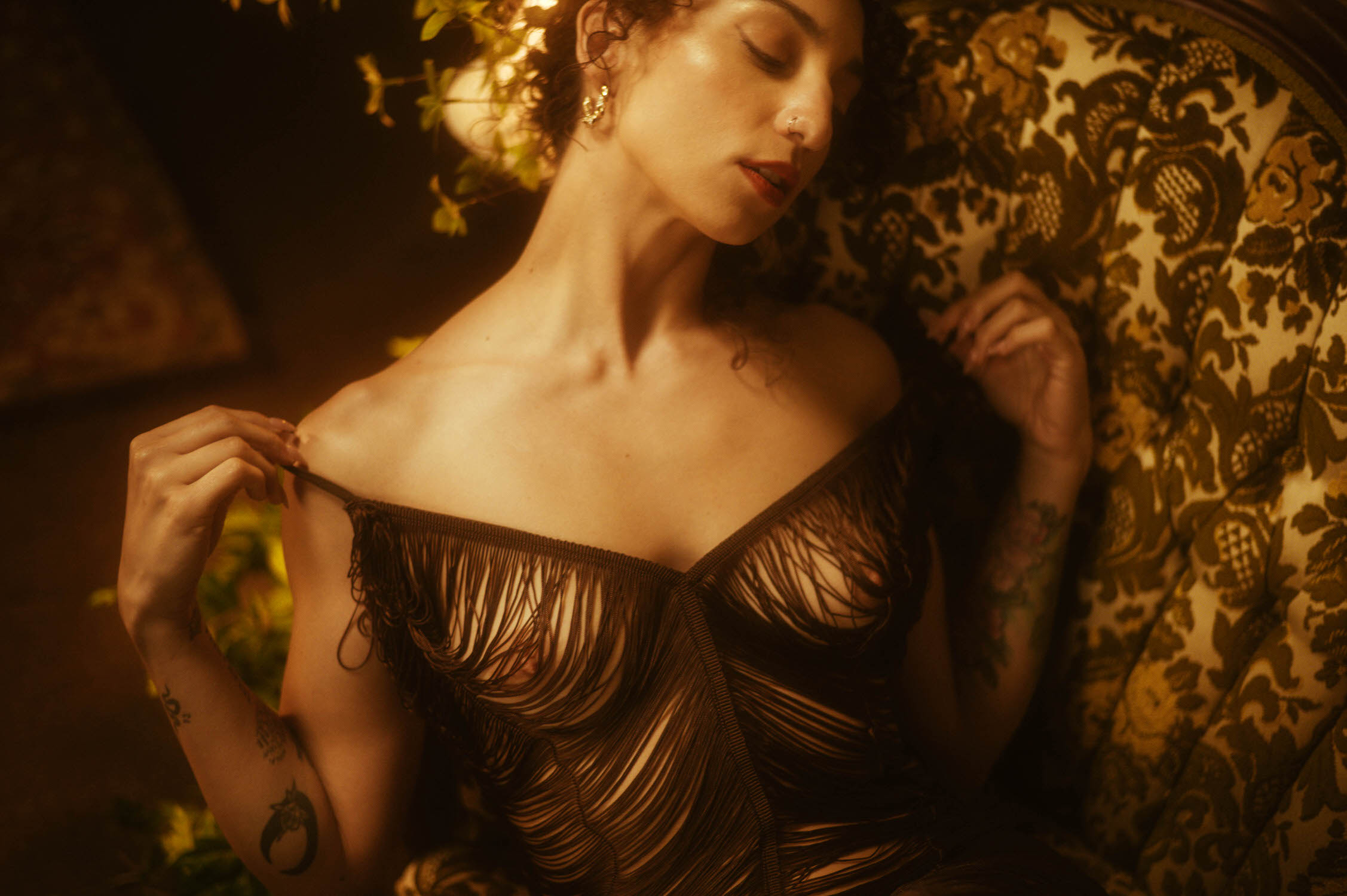 A woman with curly hair sits on a patterned chair, holding a strap of their off-the-shoulder dress. Surrounding light highlights their tattoos and golden earring.