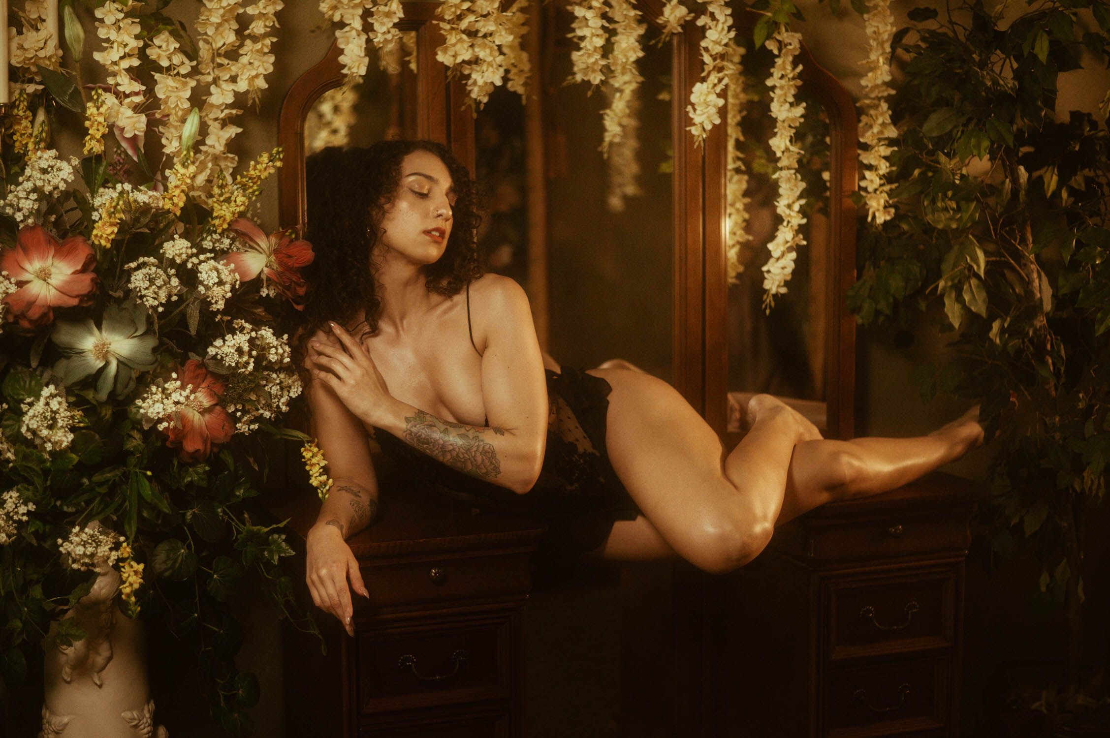 A person with curly hair in a vintage black lingerie romper lounges on a wooden vanity surrounded by flowers and greenery.