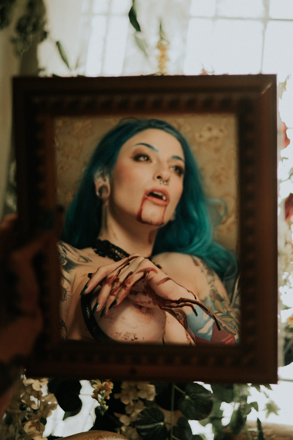 A woman with blue hair and tattoos poses with fake blood effects, holding a cloth to their chest. They are framed in a wooden picture frame surrounded by plants, capturing the sensual allure of a Halloween boudoir shoot.