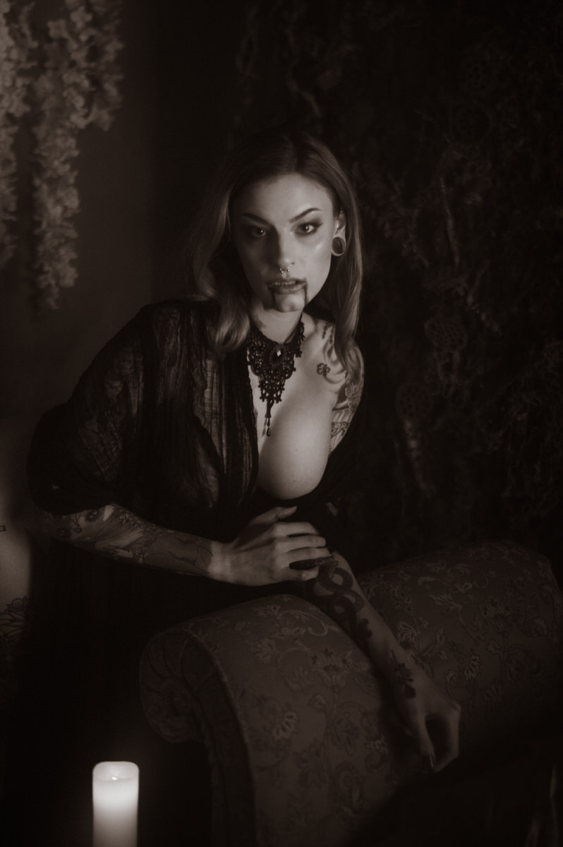 A woman dressed in dark clothing, adorned with tattoos, leans on a patterned chair in a dimly lit room beside a candle, with vines or foliage in the background—a perfect setting for a vampire photoshoot.