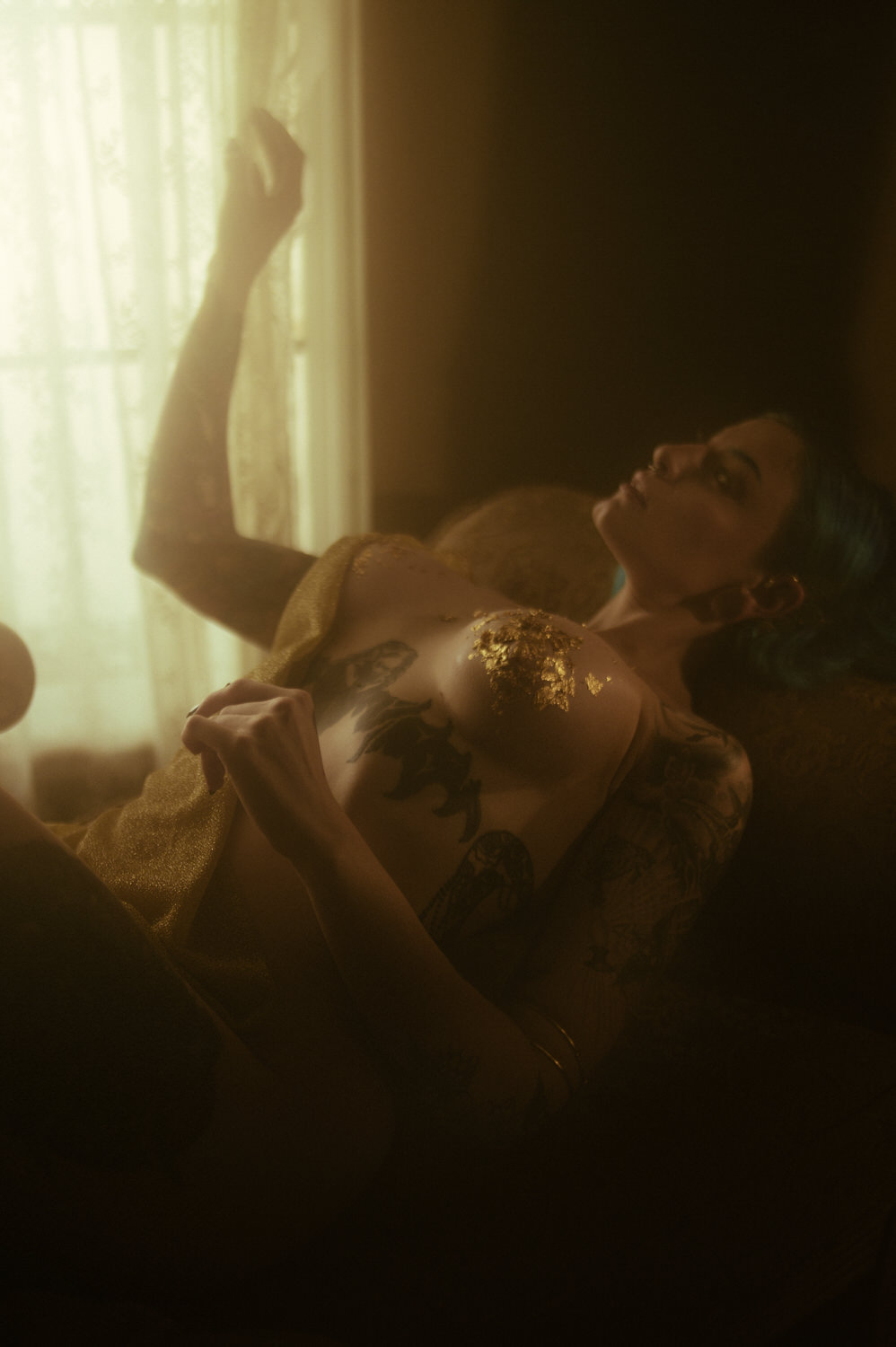 A woman with tattoos and gold leaf on their chest reclines on a couch, looking towards the light coming through sheer curtains, embodying the essence of a Dark and Ethereal, Magical Fall Boudoir Shoot.