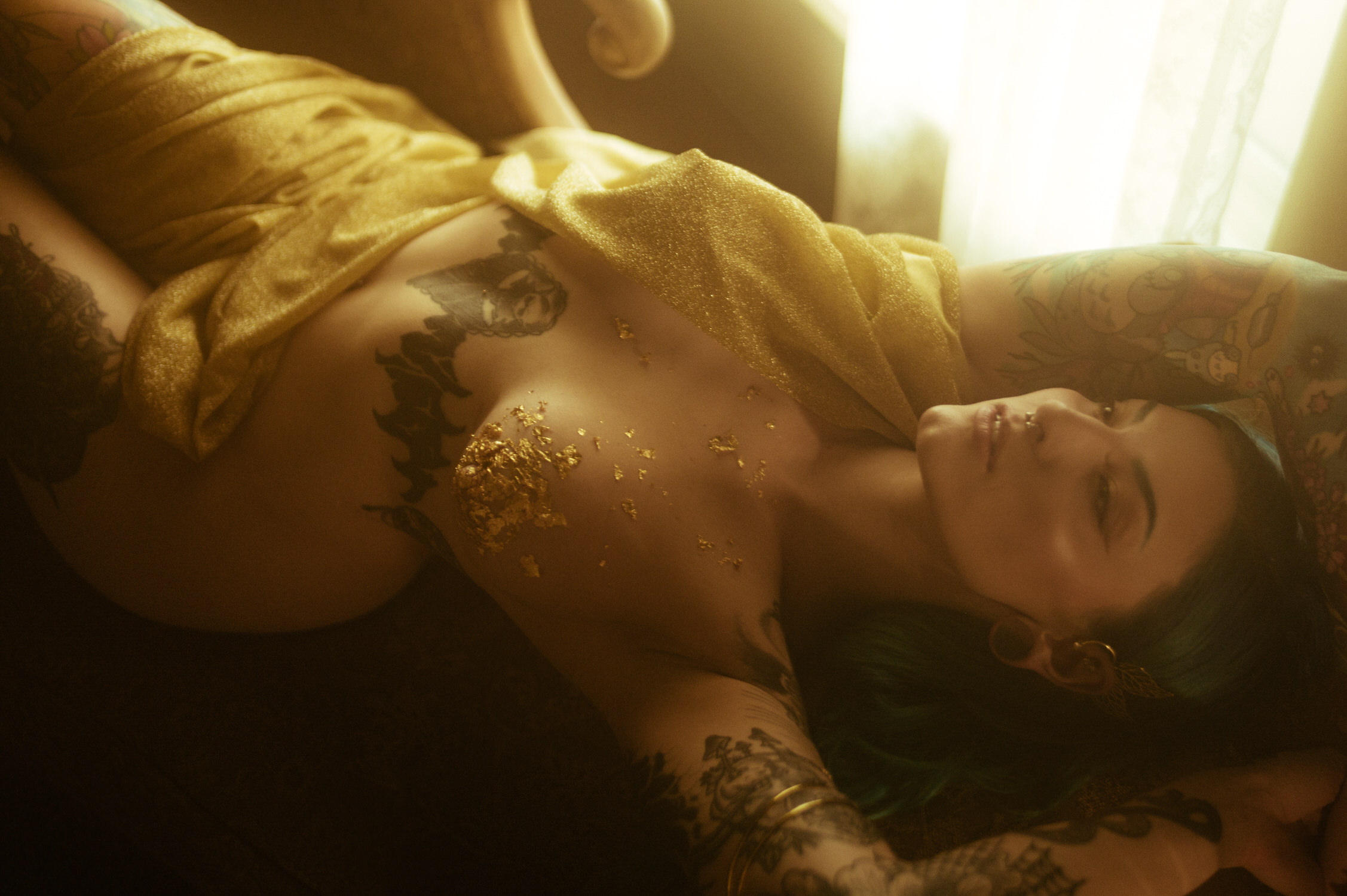 A woman with green hair, elaborate tattoos, and gold leaf on their chest, draped in yellow fabric, lying on a couch in soft lighting. The atmosphere is dark and ethereal, evoking the moody essence of a Fall Boudoir Shoot.