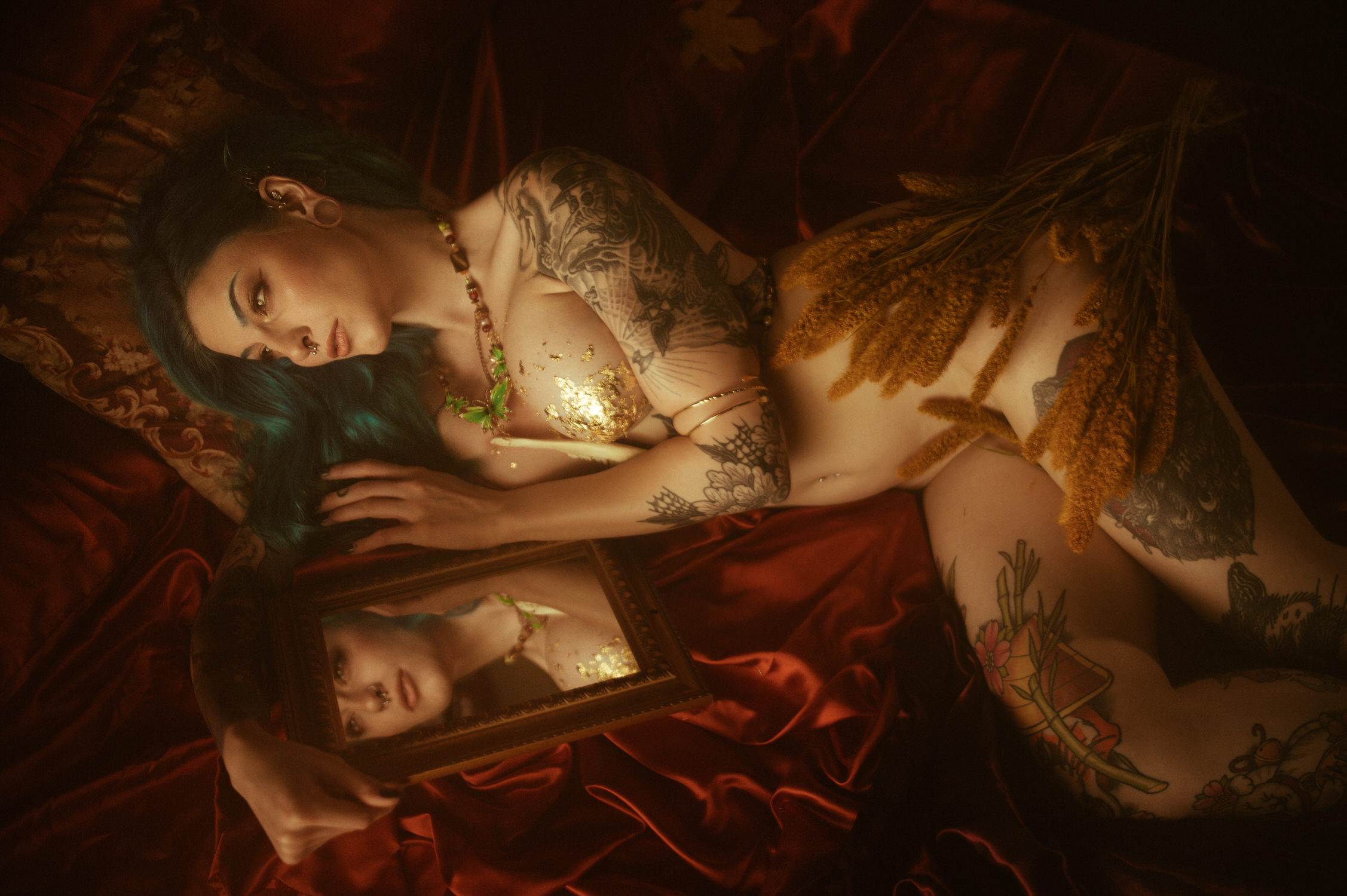 A woman with tattoos and blue hair lies on a red satin sheet in a dark and ethereal setting, holding a framed mirror reflecting their face, with dried plants placed on their body for a fall boudoir shoot in Dallas.