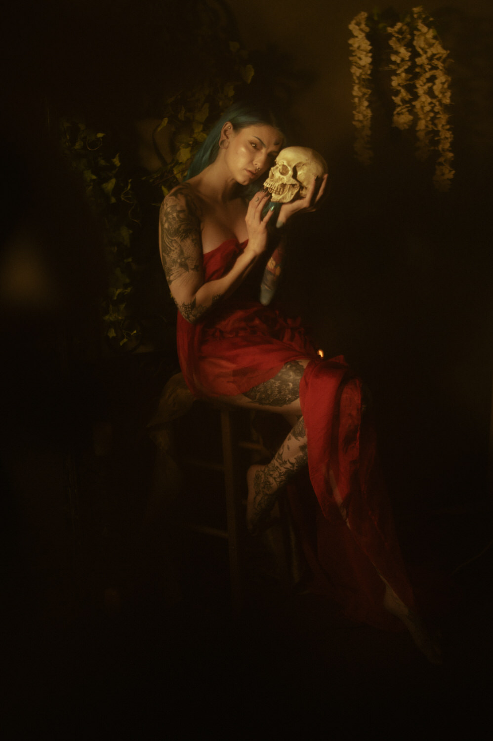 A woman with tattoos sits on a stool, wearing a red dress and holding a skull close to their face, in what looks like a Halloween boudoir photoshoot, capturing the dimly lit and mysterious environment perfectly.