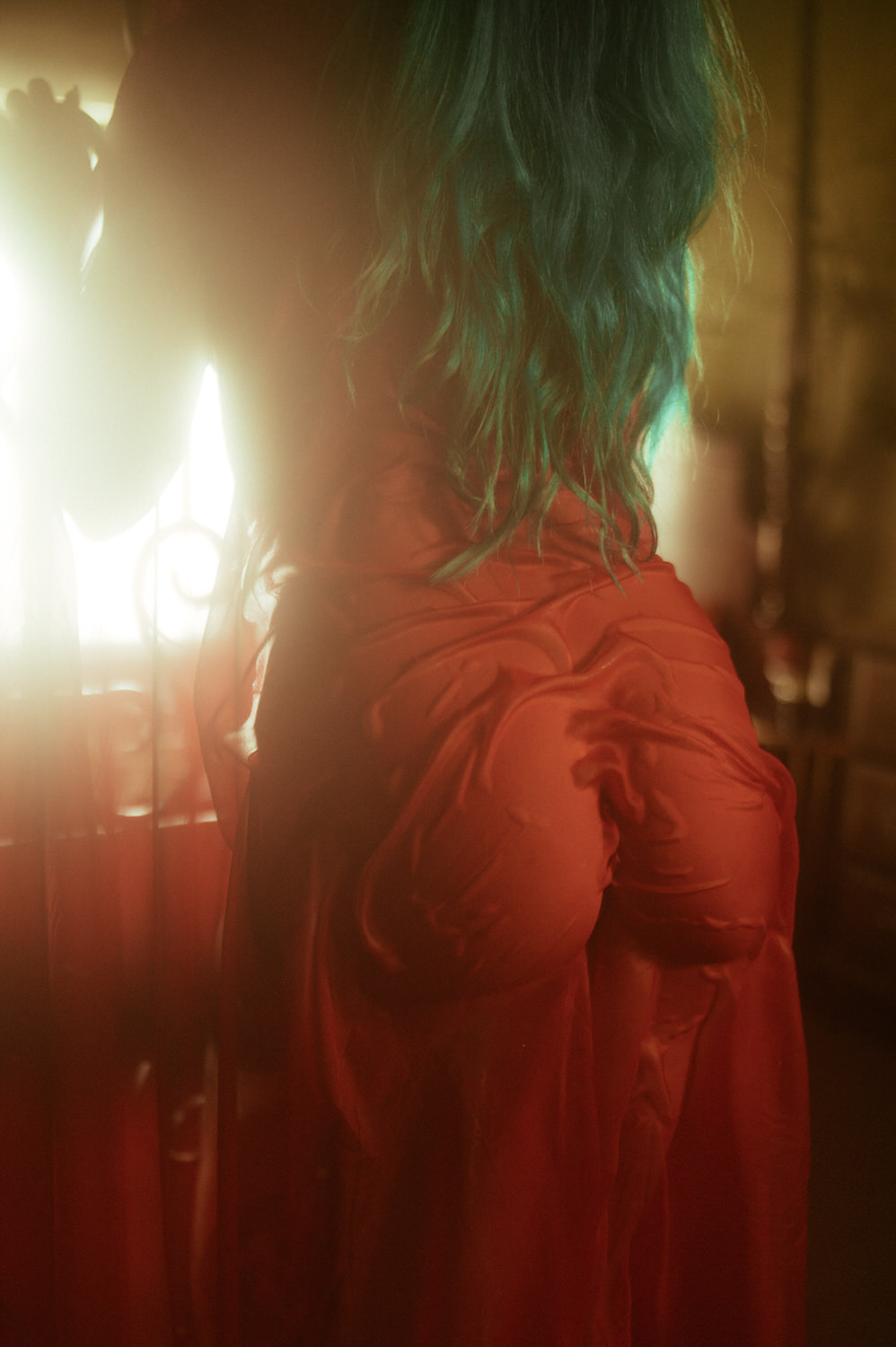 A woman with teal hair is standing with their back to the camera, wrapped in a flowing, translucent red fabric. The image has a soft, glowing light effect, perfectly capturing the essence of a mesmerizing fall boudoir shoot.