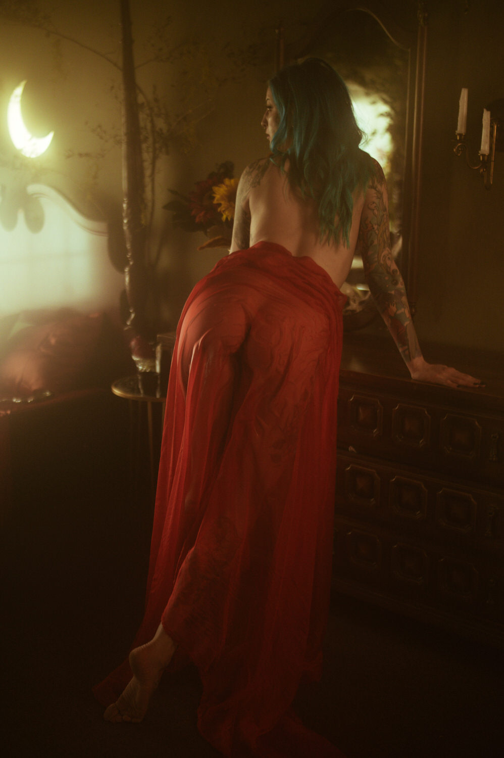 A woman with blue hair is standing beside a dresser, partially draped in a sheer wet red fabric, illuminated by soft, warm lighting. A crescent moon decoration is visible on the wall in the background, creating a dreamy atmosphere perfect for a Dallas boudoir shoot.