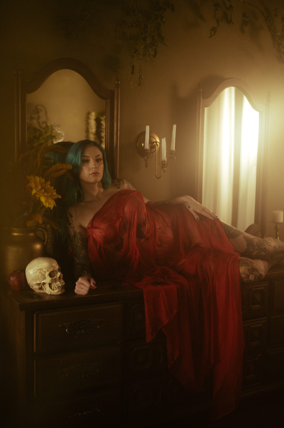 A woman with blue-green hair and tattoos reclines on a wooden dresser, draped in red fabric. A skull and small candle holder rest beside them, adding a touch of mystery to this Halloween boudoir. The room has earthy tones and soft lighting.