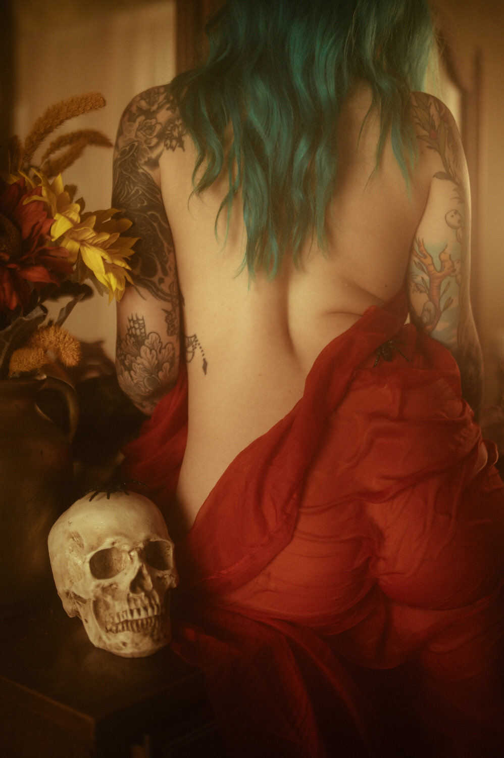 A woman with blue hair and numerous tattoos sits with their back to the camera, partially covered by a red cloth. Nearby, a skull and a vase of flowers add an eerie touch to this Halloween boudoir scene.