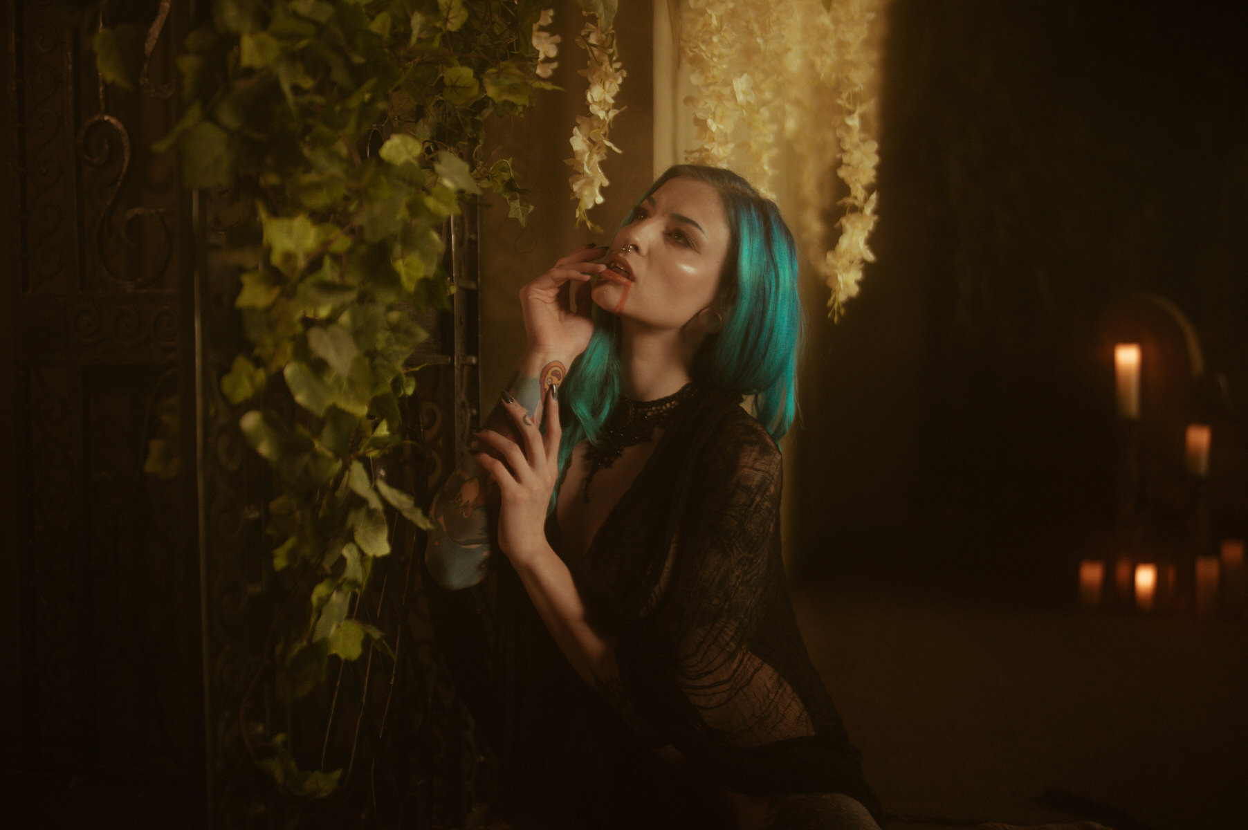 A woman with blue hair, dressed in black, kneels beside a plant-covered gate indoors, gently touching her face during a vampire themed Fall Boudoir Shoot. Candles are visible in the background.