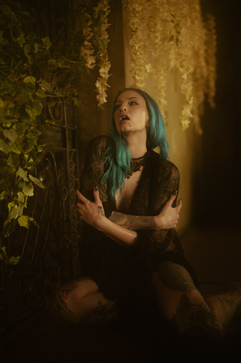 A woman with blue hair and tattoos sits on the ground, wearing a dark lace outfit, surrounded by greenery and hanging vines in dim lighting—creating a Dallas boudoir vibe perfect for a mysterious fall photoshoot.