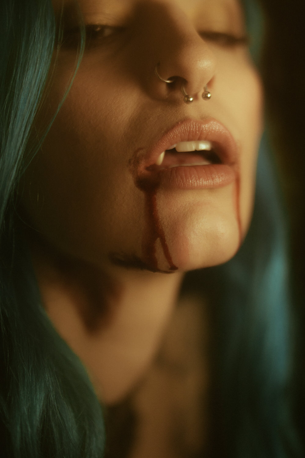 Close-up of a woman with blue hair and facial piercings, blood trickling from their mouth and chin. The dimly lit scene hints at a vampire photoshoot, creating a dramatic effect.