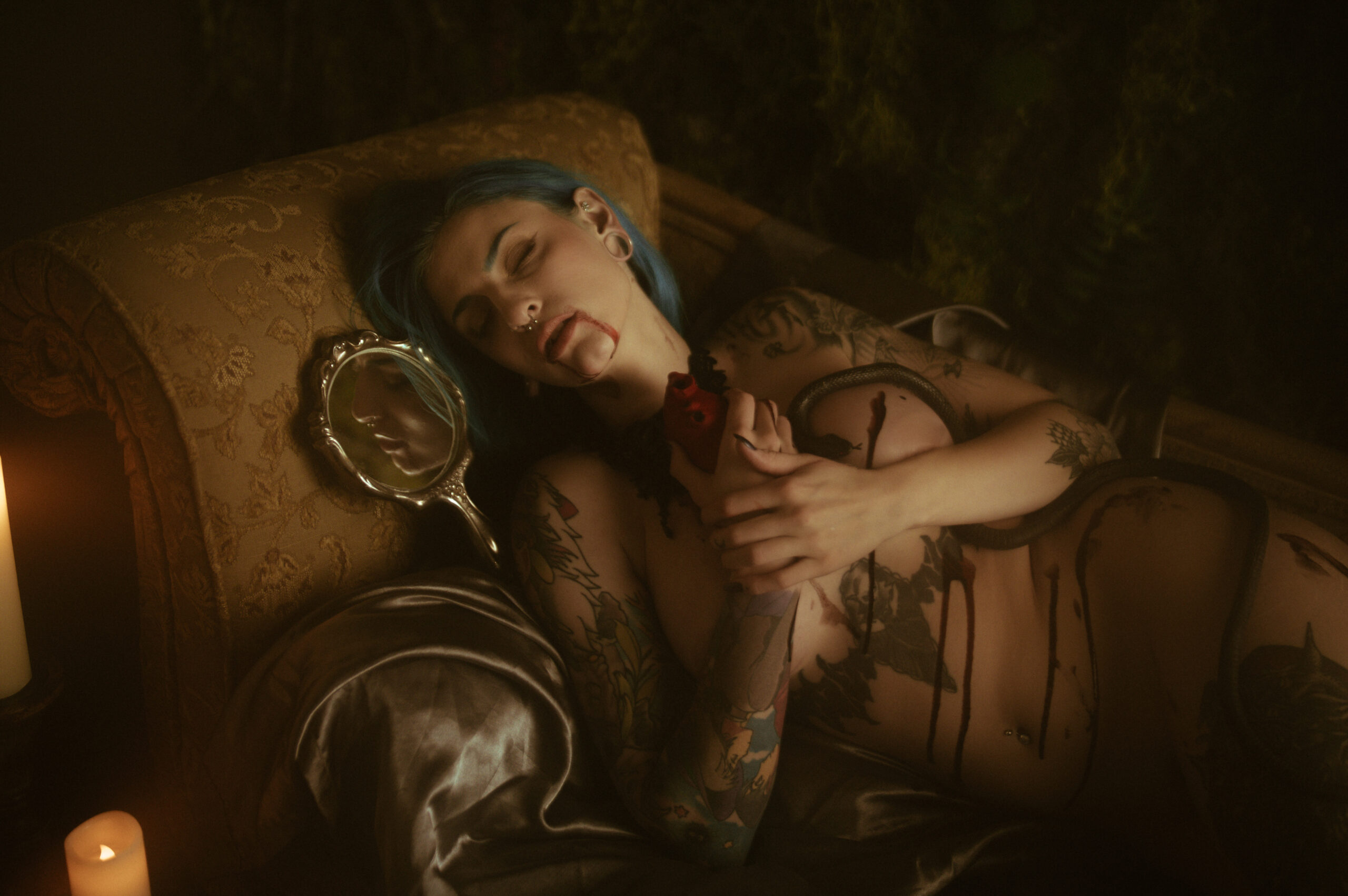 A woman with blue hair and body tattoos dressed as a vampire lies on a couch holding a bloody heart. A lit candle and a hand mirror are beside them, creating an ethereal atmosphere for this fall boudoir shoot.