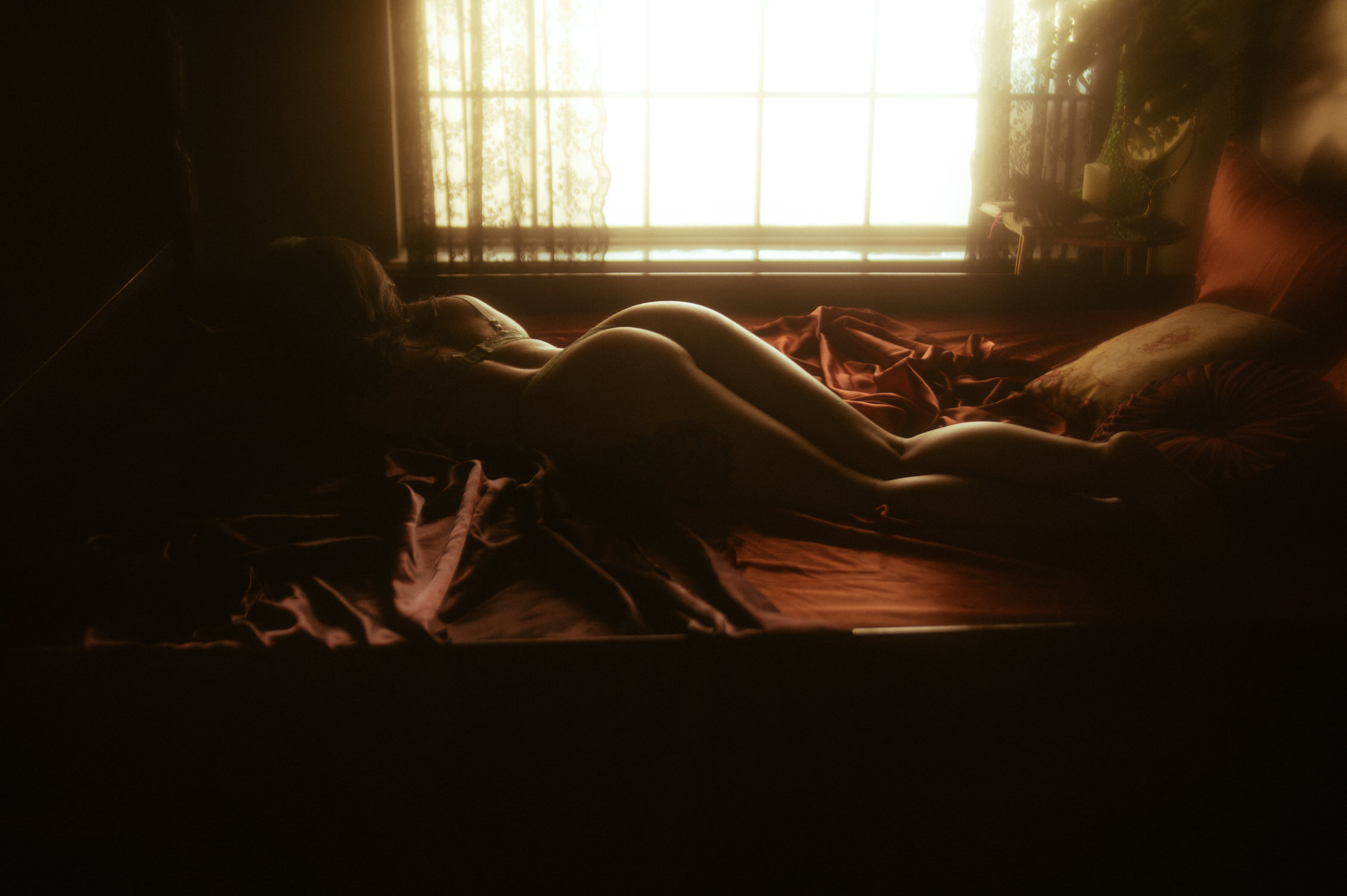 A woman lying nude face down on a bed with red sheets, next to a window with light filtering through sheer curtains, creates the perfect scene for a Dallas boudoir session.