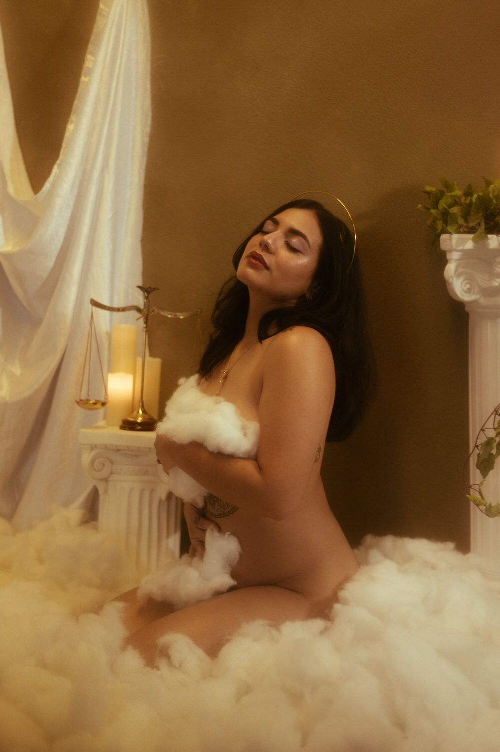 A woman poses kneeling on a soft white surface, surrounded by cotton-like material, with candles, a balance scale, and a Greek column nearby in this ethereal Libra photoshoot.