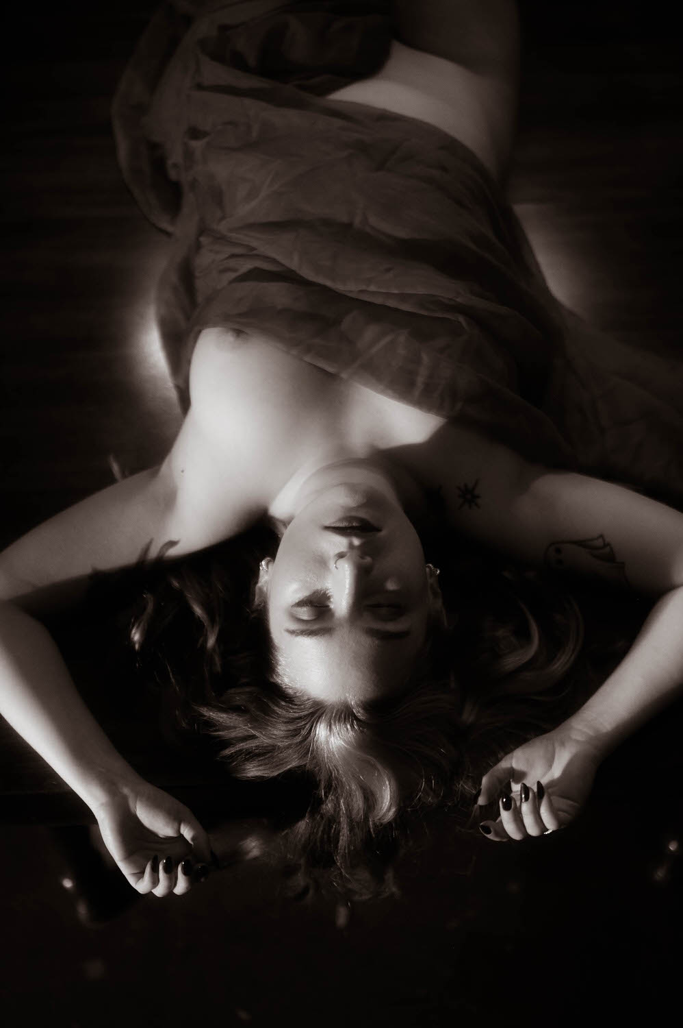 A woman lies down with eyes closed, partially covered by a draped cloth in a dimly lit setting, reminiscent of NYC Boudoir Photography. Arms are placed beside the head with visible tattoos on shoulders, evoking a subtle witchy boudoir vibe.