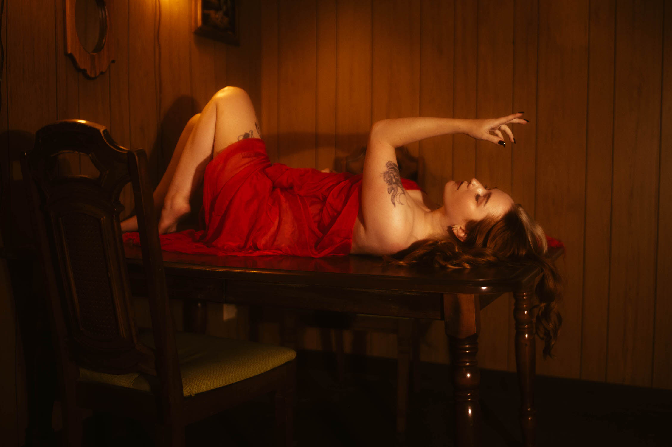 A woman lying on a wooden table, partially covered by a red cloth, in a dimly lit room with wood-paneled walls, creating an atmosphere perfect for this NYC boudoir photography session with witchy themes. The scene is spellbinding, evoking an aura of mystery and allure 