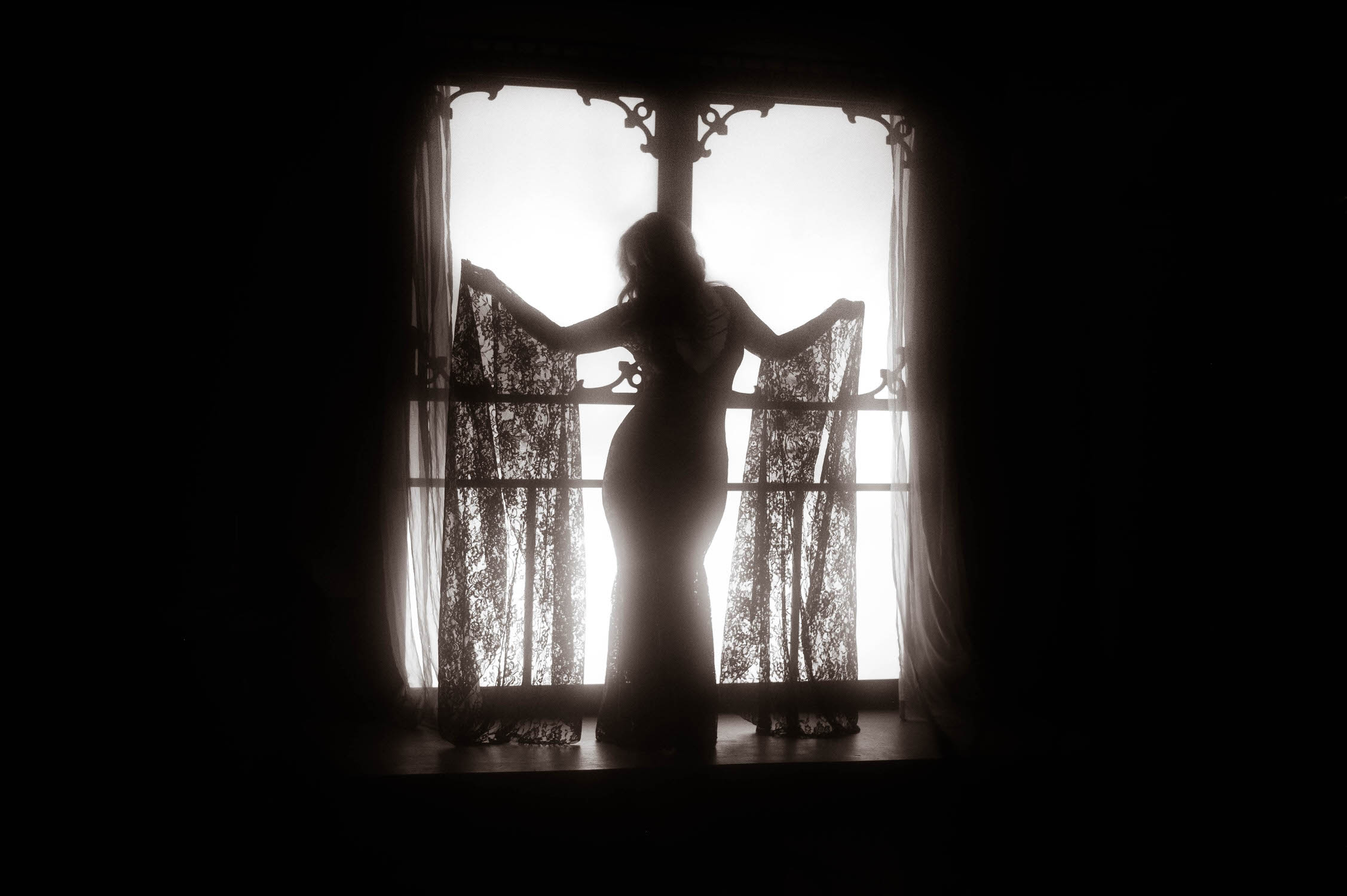 A silhouette of a woman in a gothic lace dress standing at a window, with light streaming in from behind, evoking an enchanting Witchy Boudoir vibe in New York City.