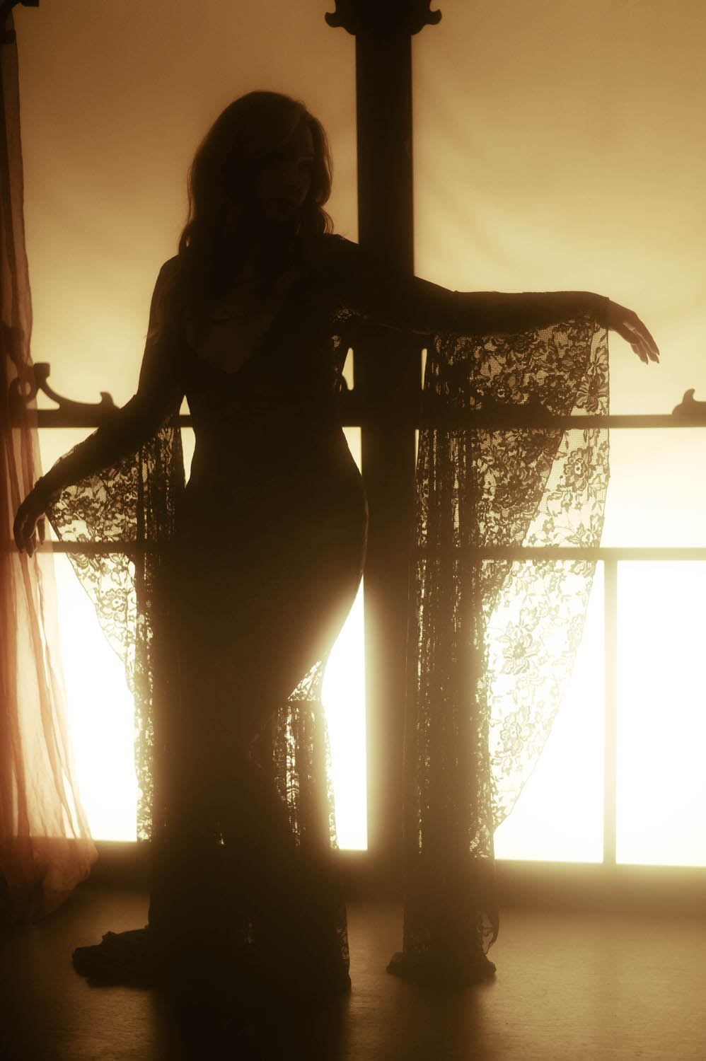 A silhouette of a woman in a lace dress standing against a brightly lit window with arms extended, exuding a spellbinding presence.