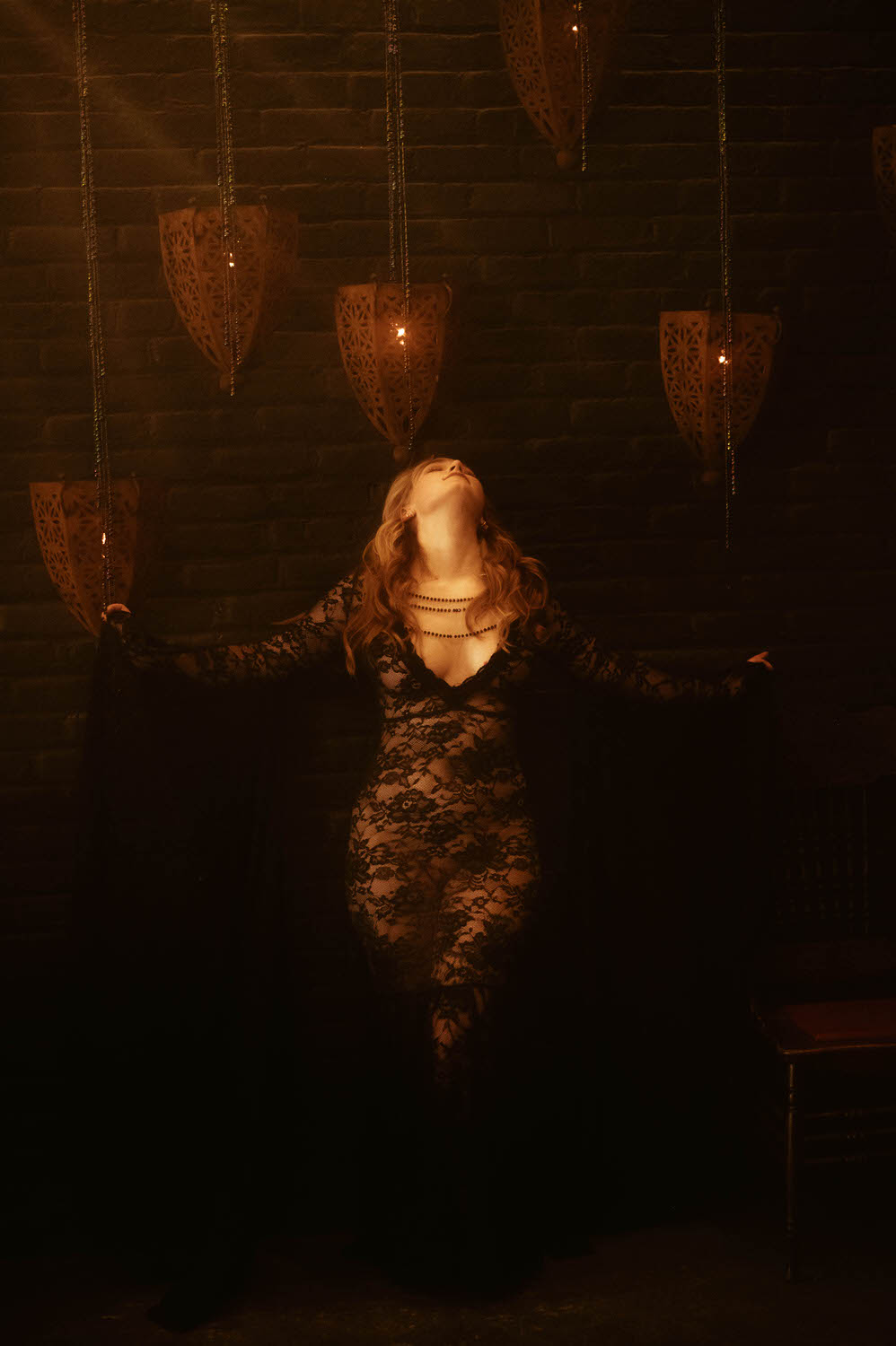 A woman wearing a long black lace dress stands with arms extended in front of a dark brick wall adorned with hanging lanterns, evoking a witchy boudoir vibe.