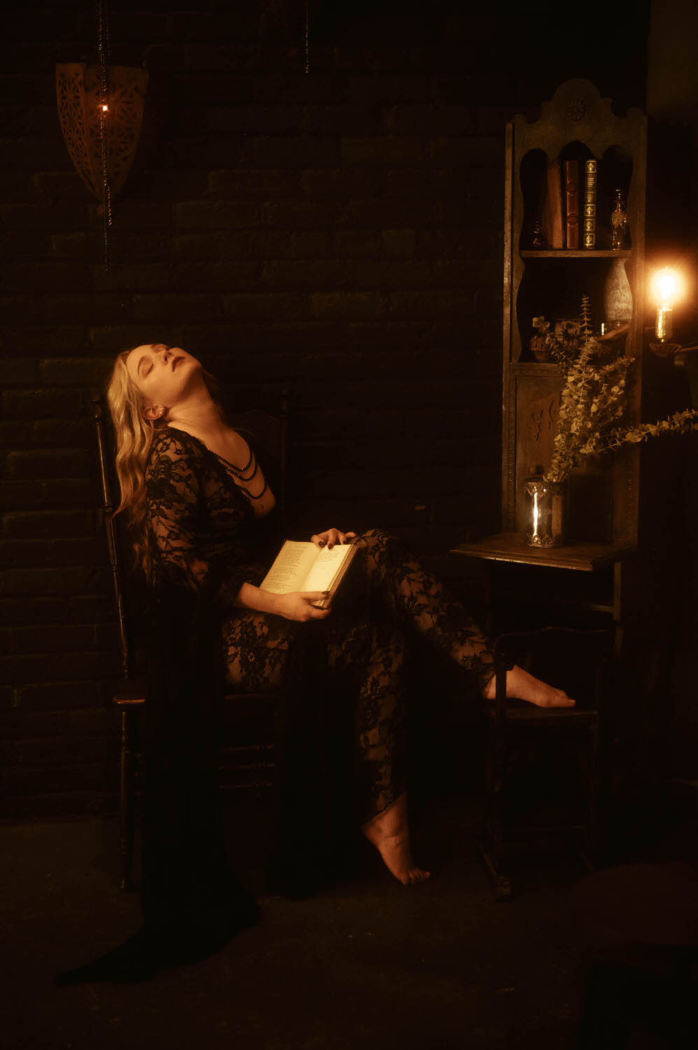 A woman with long blonde hair sits on a wooden chair in a dimly lit room with brick walls, holding an open book and leaning back with their eyes closed. A candle and wooden shelves are beside them, creating a scene akin to an NYC Boudoir Photography session.