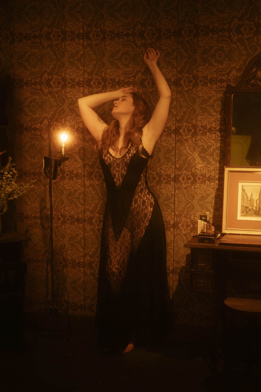 A woman stands in a dimly lit room with one arm raised, wearing a black, sheer lace dress. She poses against ornate wallpaper beside a lit wall sconce and a desk with framed art, embodying the enchanting allure of Witchy Boudoir in NYC.