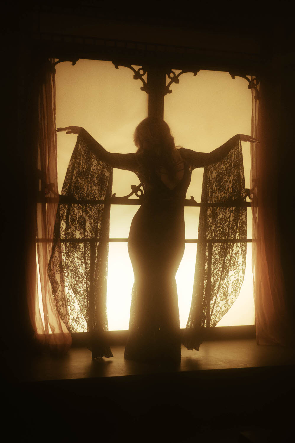 A woman in a long dress with lace sleeves poses in front of an illuminated window with curtains, their silhouette creating a spellbinding scene against the light for a NYC boudoir photography session.