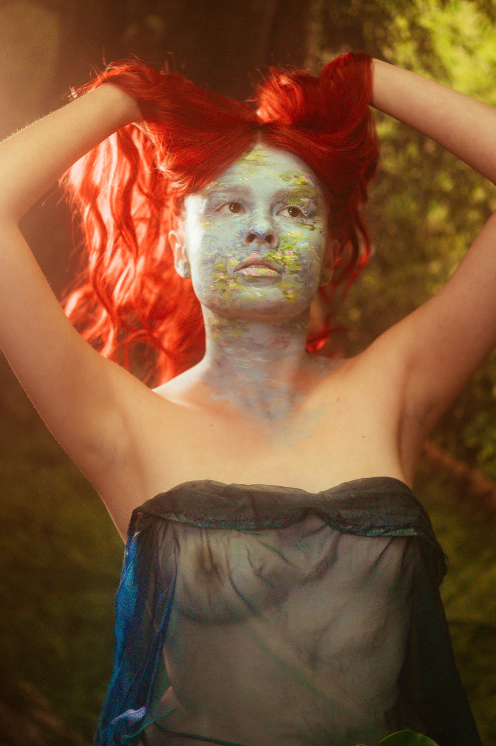 A woman with bright red hair, adorned in striking blue body paint, stands confidently with arms raised against a nature-themed backdrop, blending elegance and wild allure.