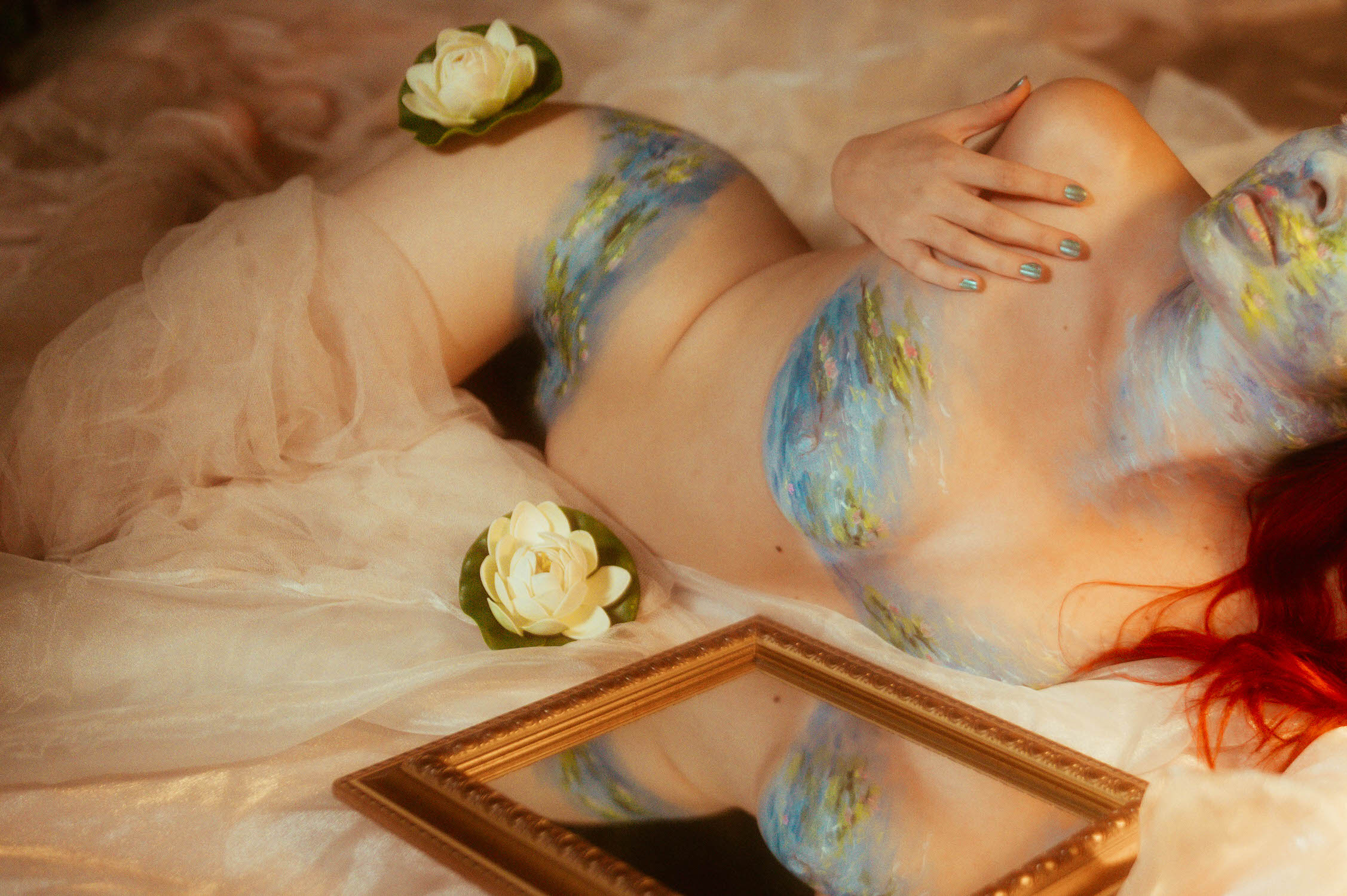 A woman with body paint lies on a fabric surface, surrounded by flowers and a mirror, capturing the essence of an artistic body paint photoshoot.