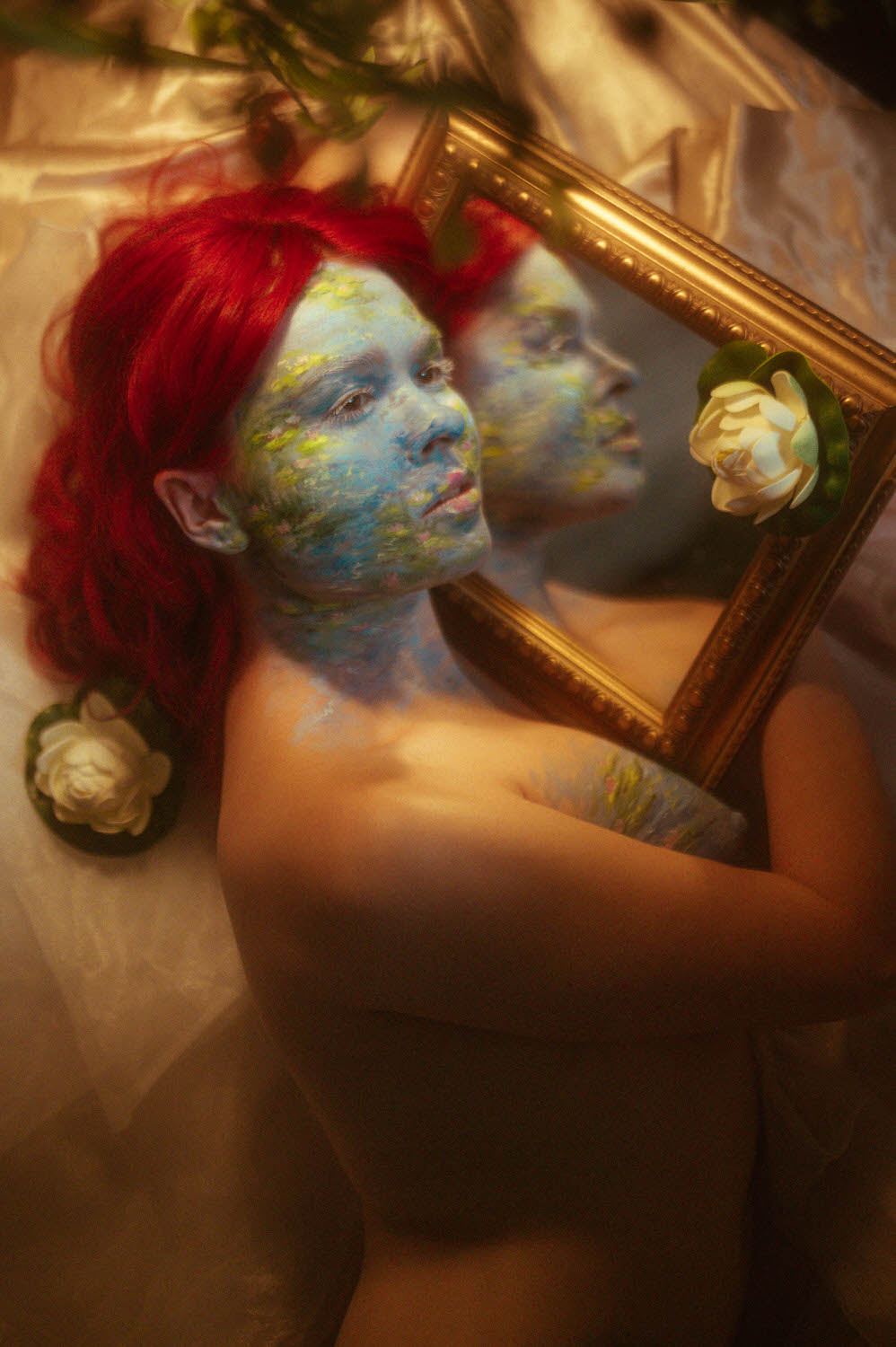 A woman with red hair, covered in vibrant body paint, holds a framed mirror reflecting their image. The scene, reminiscent of Dallas body paint photography, is set on soft, silky fabric with white flowers adorning the frame.