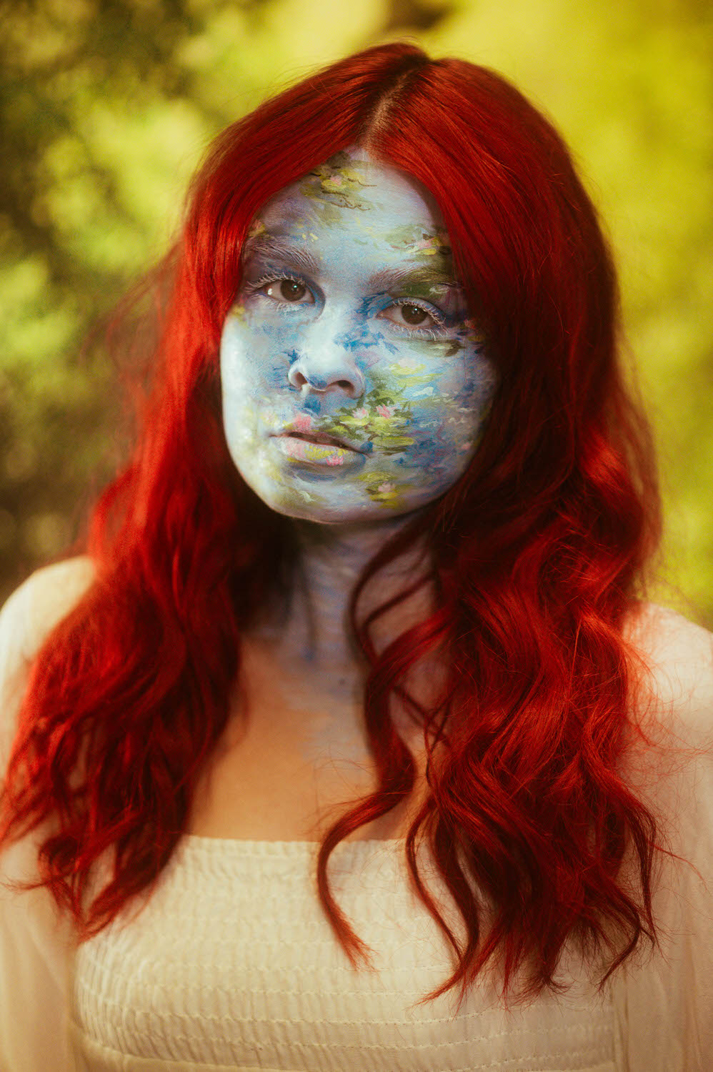 A woman with vibrant red hair, wearing white, stands against a blurred green background, their face and neck adorned with Monet-inspired body paint in blue and green earth-like designs. This enchanting photoshoot captures the essence of art and nature merging beautifully.