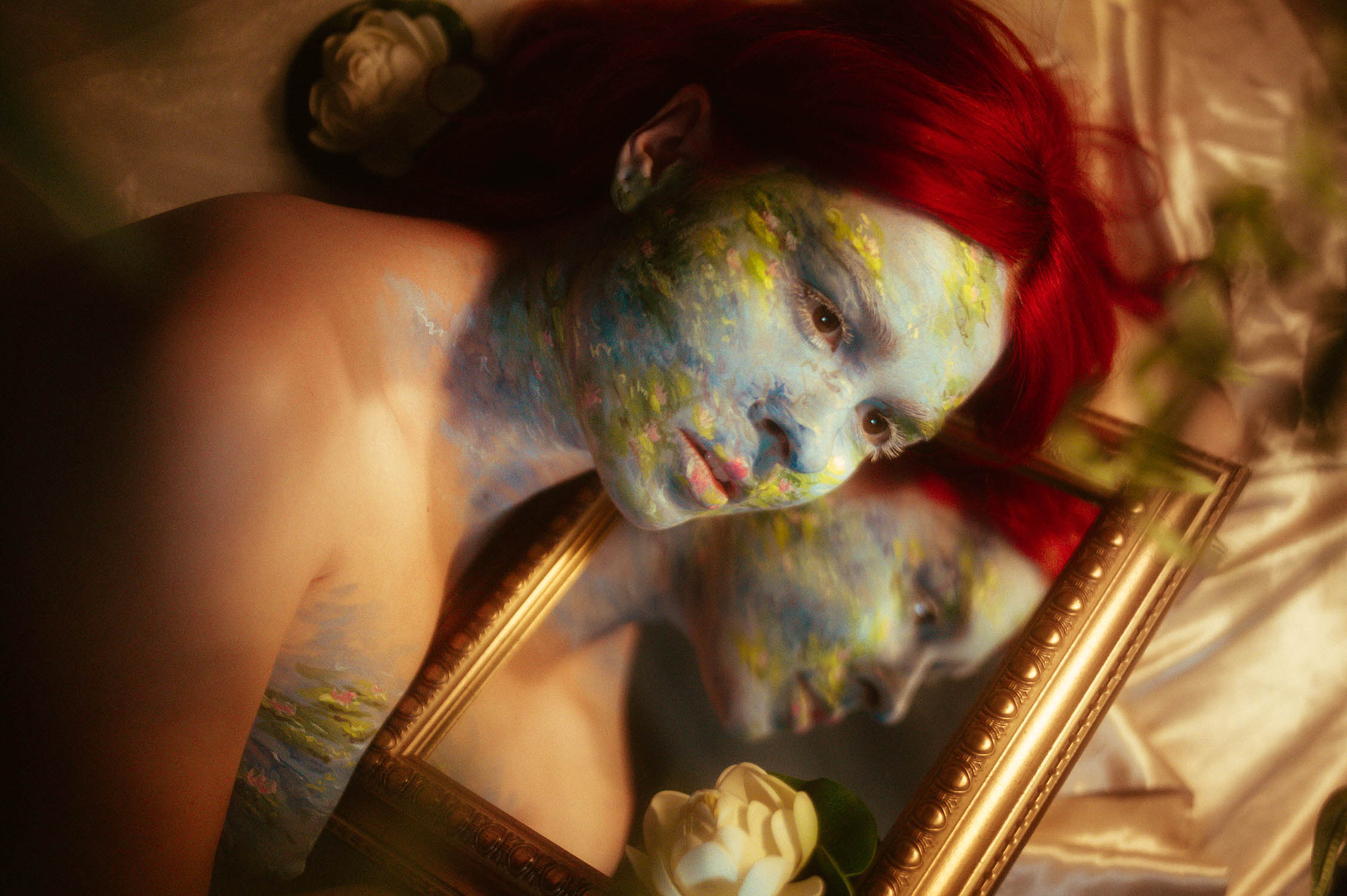 The woman with red hair and blue-green painted face, showcasing special effect makeup inspired by Monet, lies on satin fabric holding a framed mirror with their reflection. White flowers are placed around.