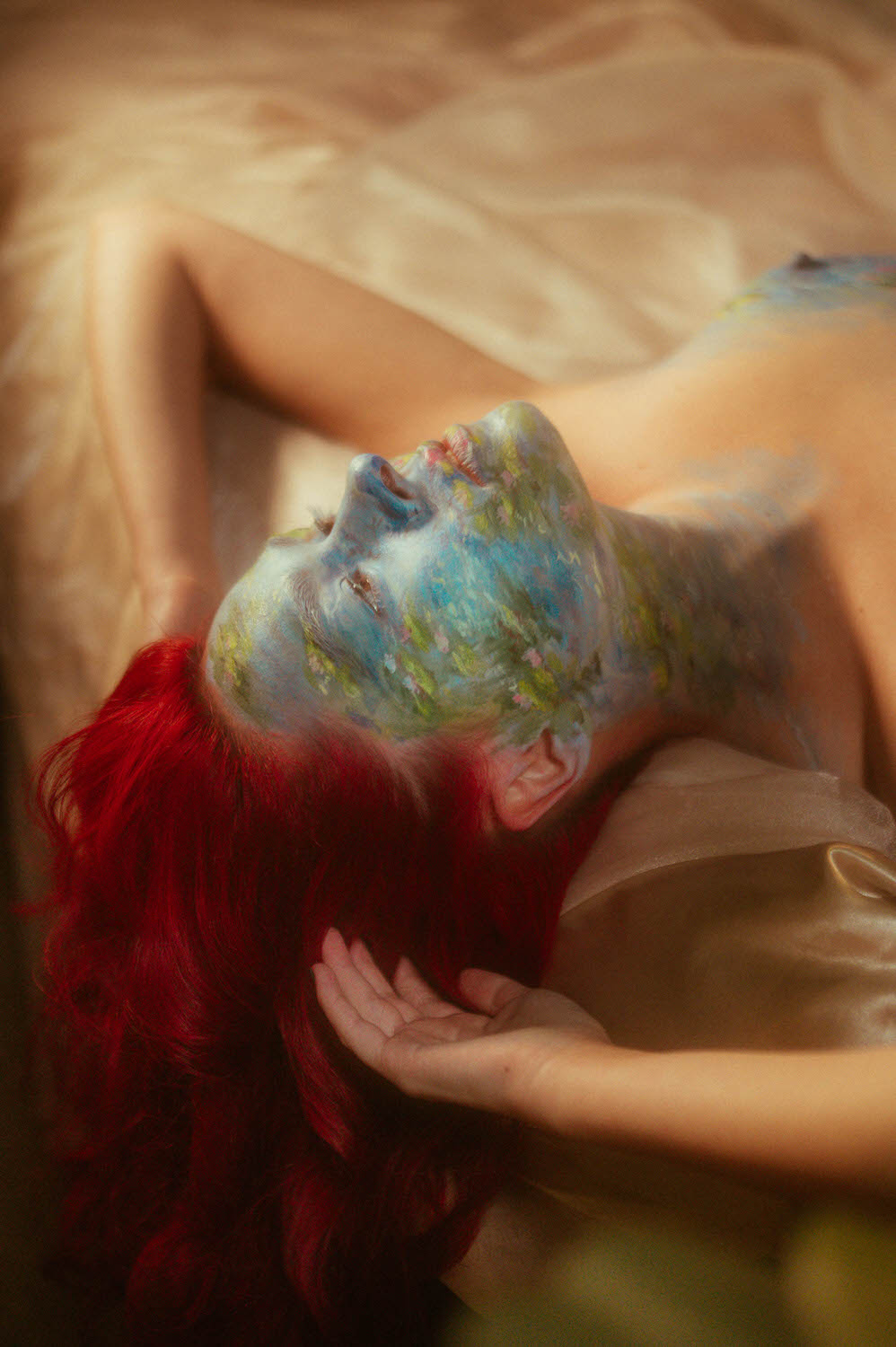 A woman with vibrant red hair lies on a beige surface, their face adorned with an abstract landscape design in stunning body paint photography.