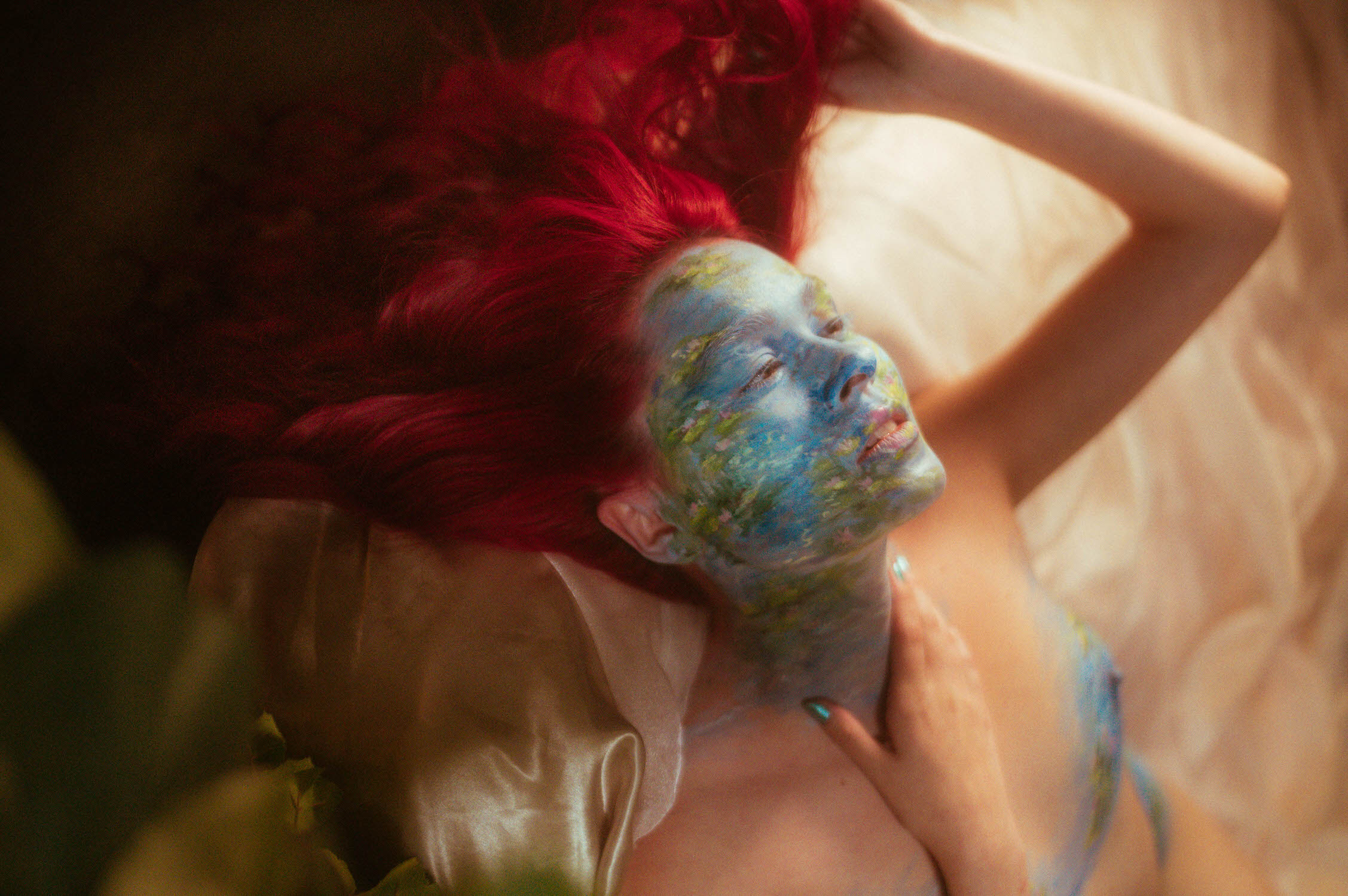 A woman with vibrant red hair lies on a satin fabric in a special effect makeup photoshoot. Their face is painted blue with green and yellow abstract patterns, one hand resting gently on their chest.