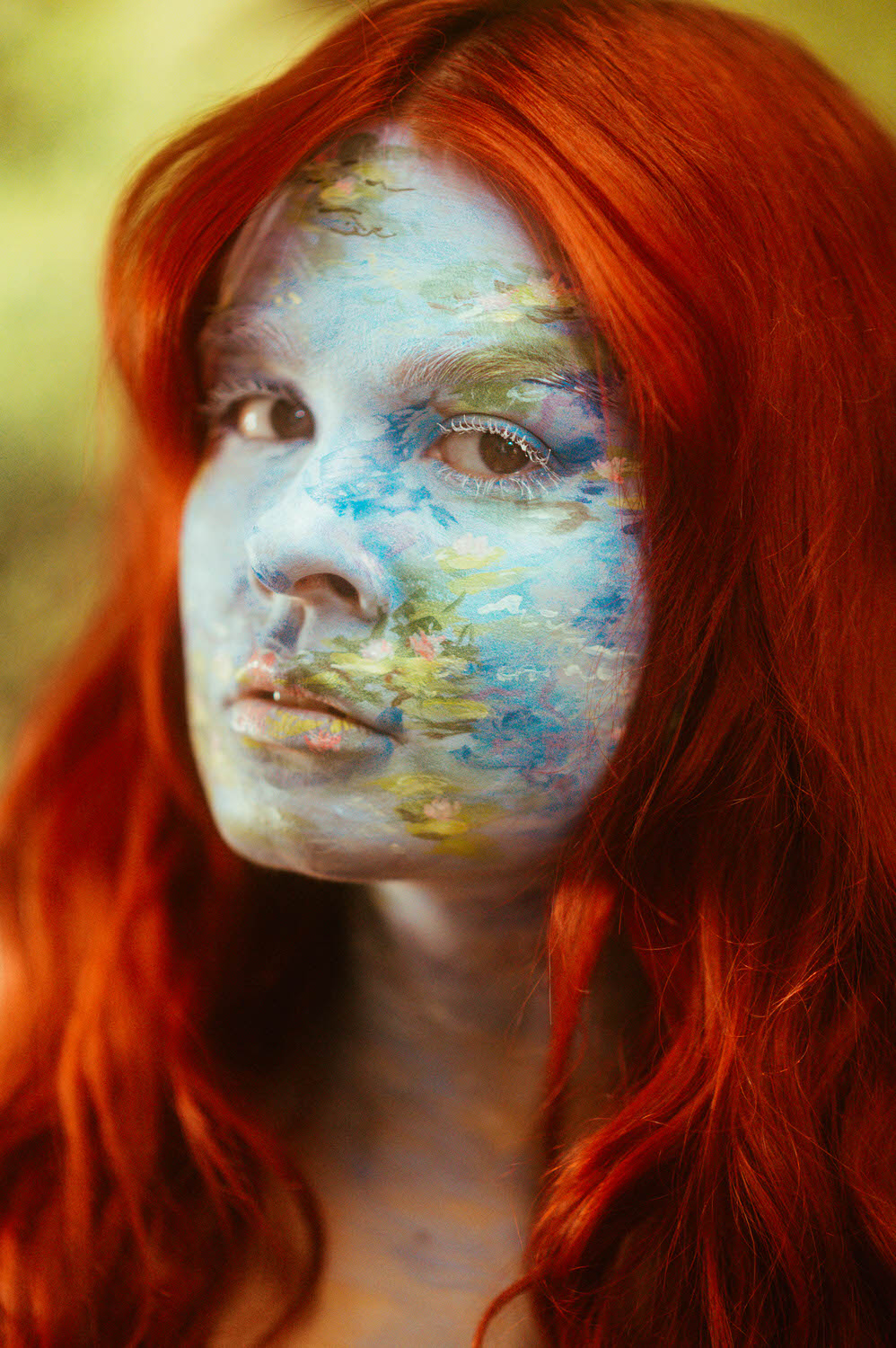 A woman with vibrant red hair stands in a captivating body paint photoshoot, their face adorned with a stunning blue and green nature scene.