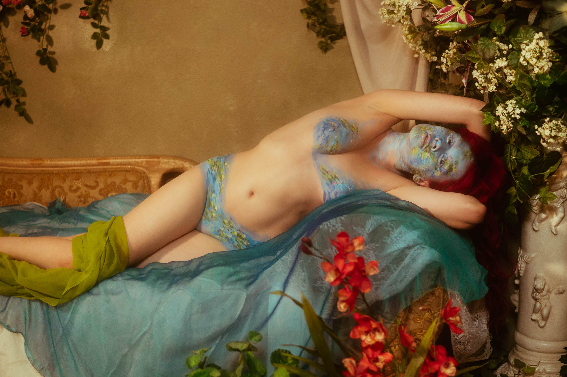 A woman with Monet-inspired body paint lies on a bed surrounded by flowers and greenery, draped with blue and green fabric, creating a serene photoshoot scene.