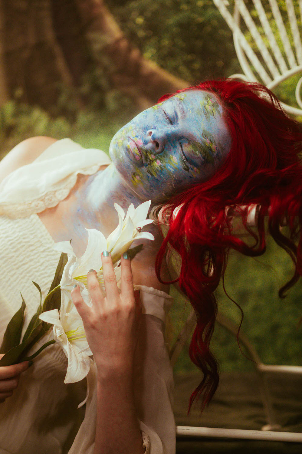 A woman with stunning special effect makeup, featuring blue face paint and red hair, holds white lilies while wearing a white garment. They lean back against a wooden structure in this serene natural setting.