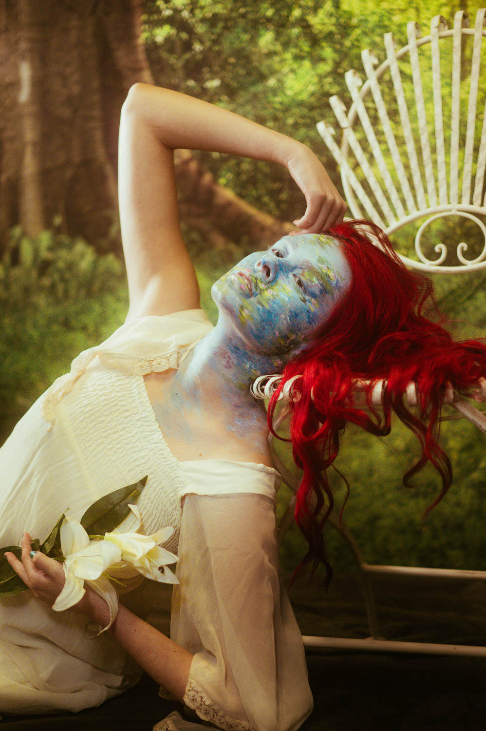 A woman with red hair poses outdoors, their face adorned in impressionist body paint featuring a nature design. They're holding white flowers and wearing a light-colored dress.