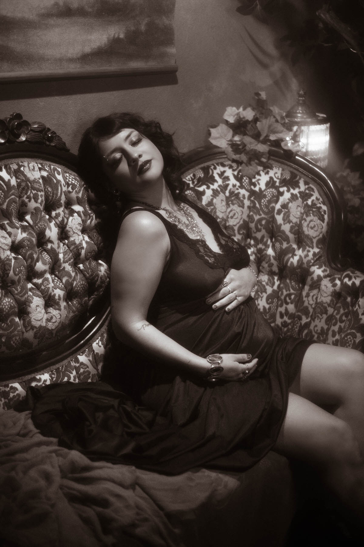 A dramatic portrait of a pregnant woman in a black lace gown, seated on an ornate vintage couch with rich, floral upholstery. She cradles her baby bump with one hand, her head tilted back, eyes closed in a serene expression, with soft, cinematic shadows adding depth to the scene. The setting includes warm, moody lighting with textured walls, evoking a timeless, romantic atmosphere.