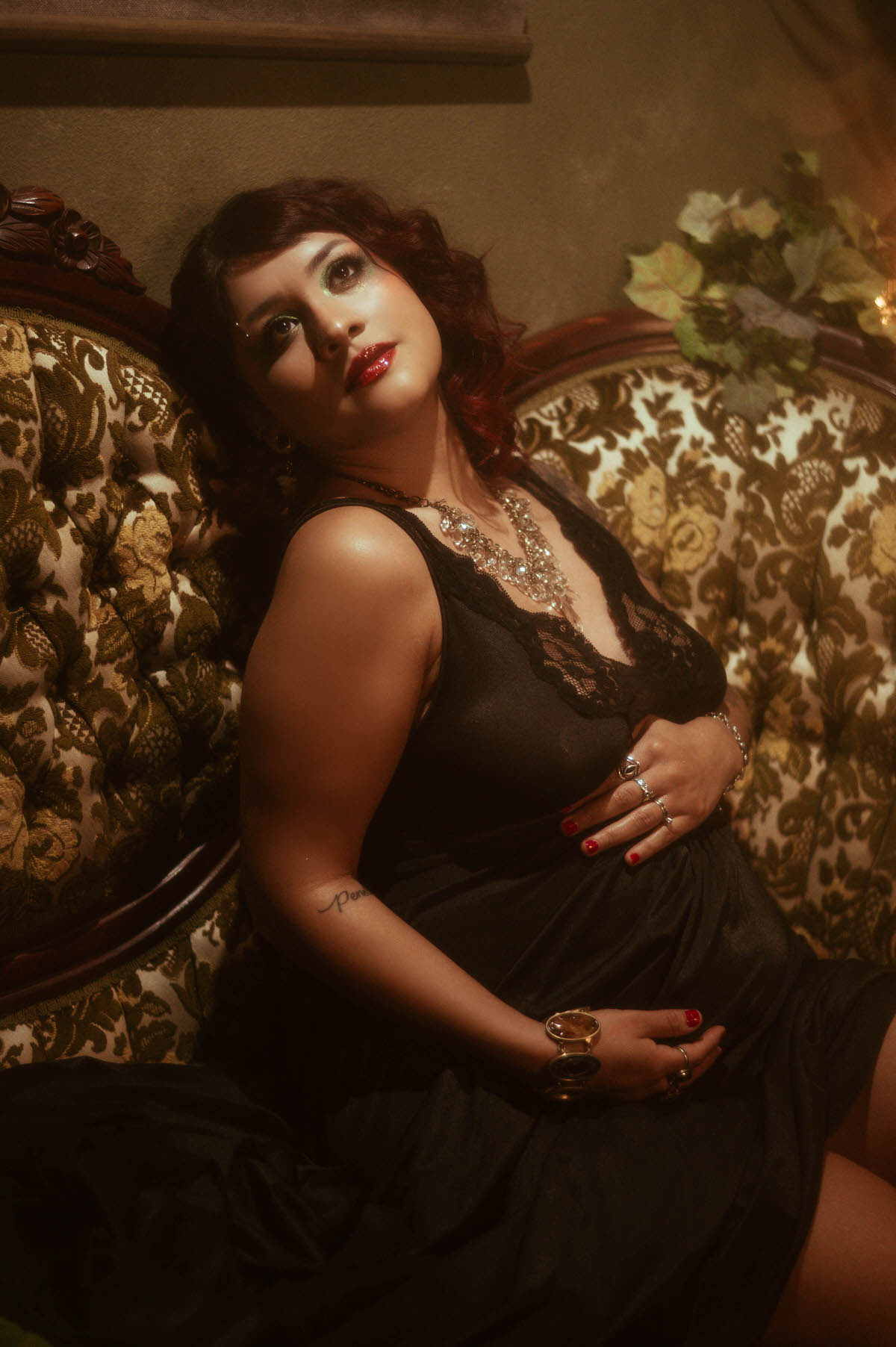 A softly lit portrait capturing a pregnant woman in a black lace dress, leaning against a vintage floral-patterned couch. Her gaze is directed upward, eyes glistening in the warm, ambient light, highlighting her glossy red lips and radiant, expectant expression. The scene is accented by lush greenery and classic antique decor, creating a rich, intimate atmosphere.
