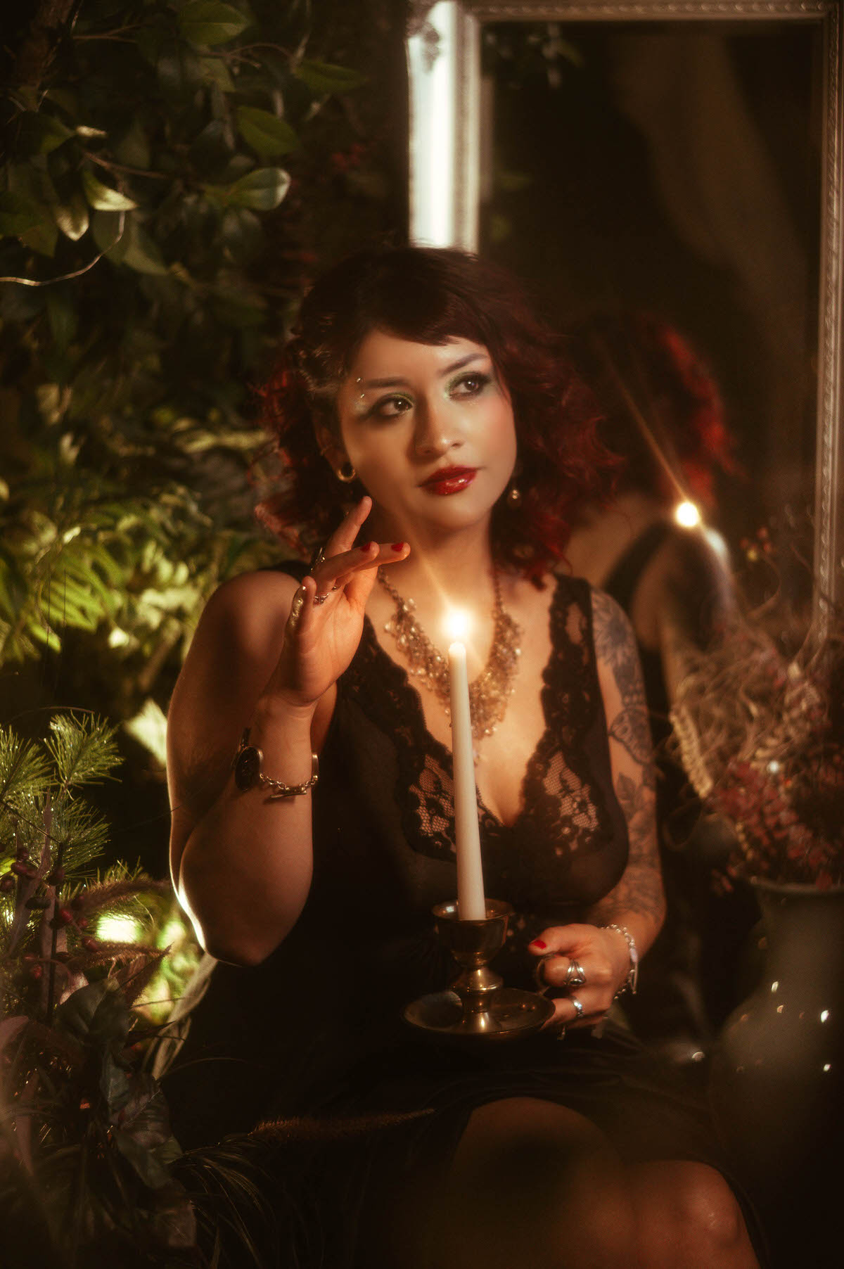 A close-up portrait of a woman in a black lace dress, holding a single tall candle in a brass holder. She gently raises her hand toward the flickering flame, her red lips and vibrant makeup catching the warm, golden light. The backdrop features dense greenery, softly blurred to frame her contemplative expression, creating a mystical, ethereal vibe.