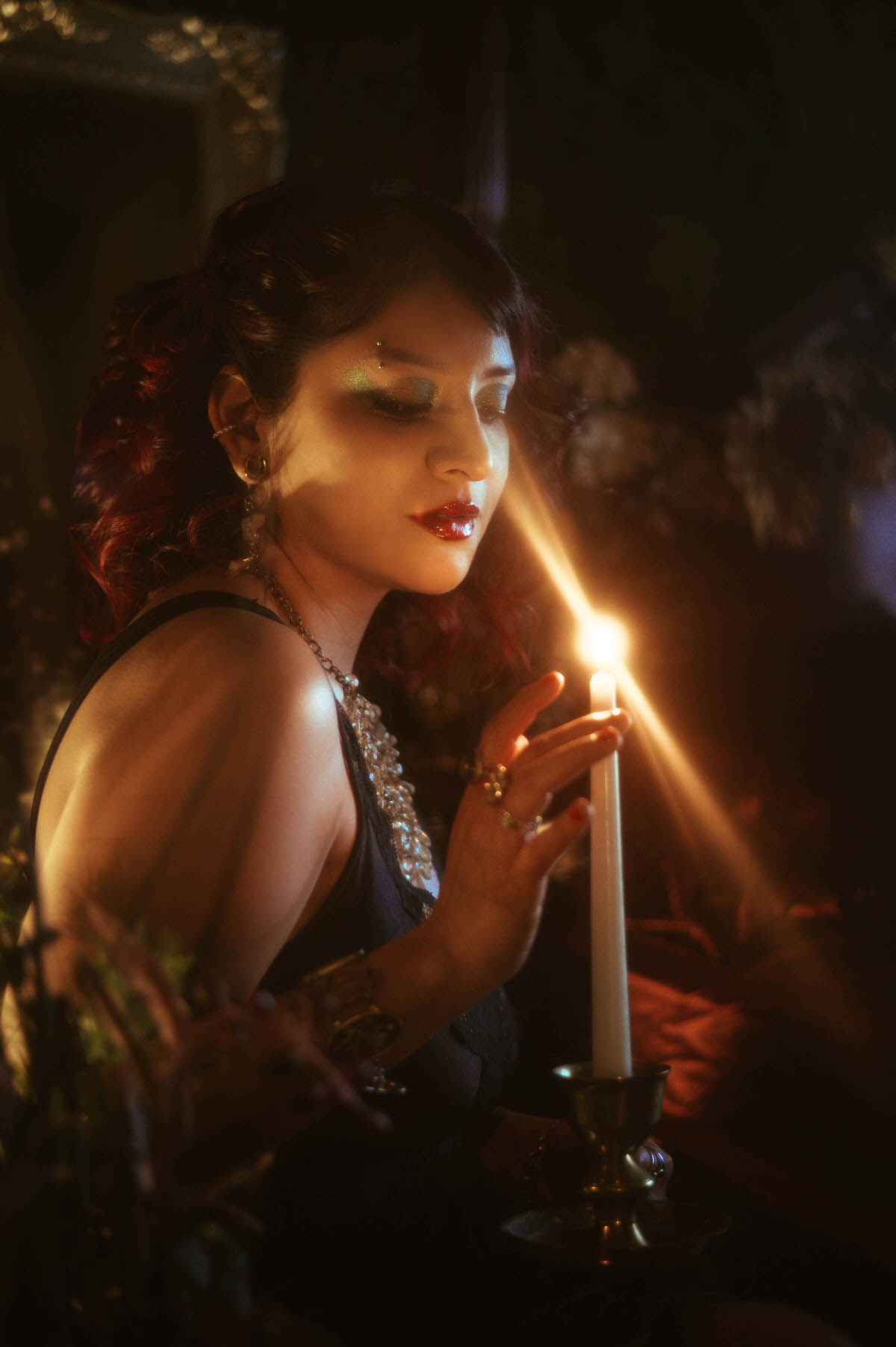 A dreamy, ethereal portrait of a pregnant woman in a black lace gown, holding a brass candlestick as she leans toward the flame. Her eyes are closed, red lips softly parted, as the warm candlelight casts a golden glow across her face, contrasting with the dark, shadowy background of lush greenery and antique decor.